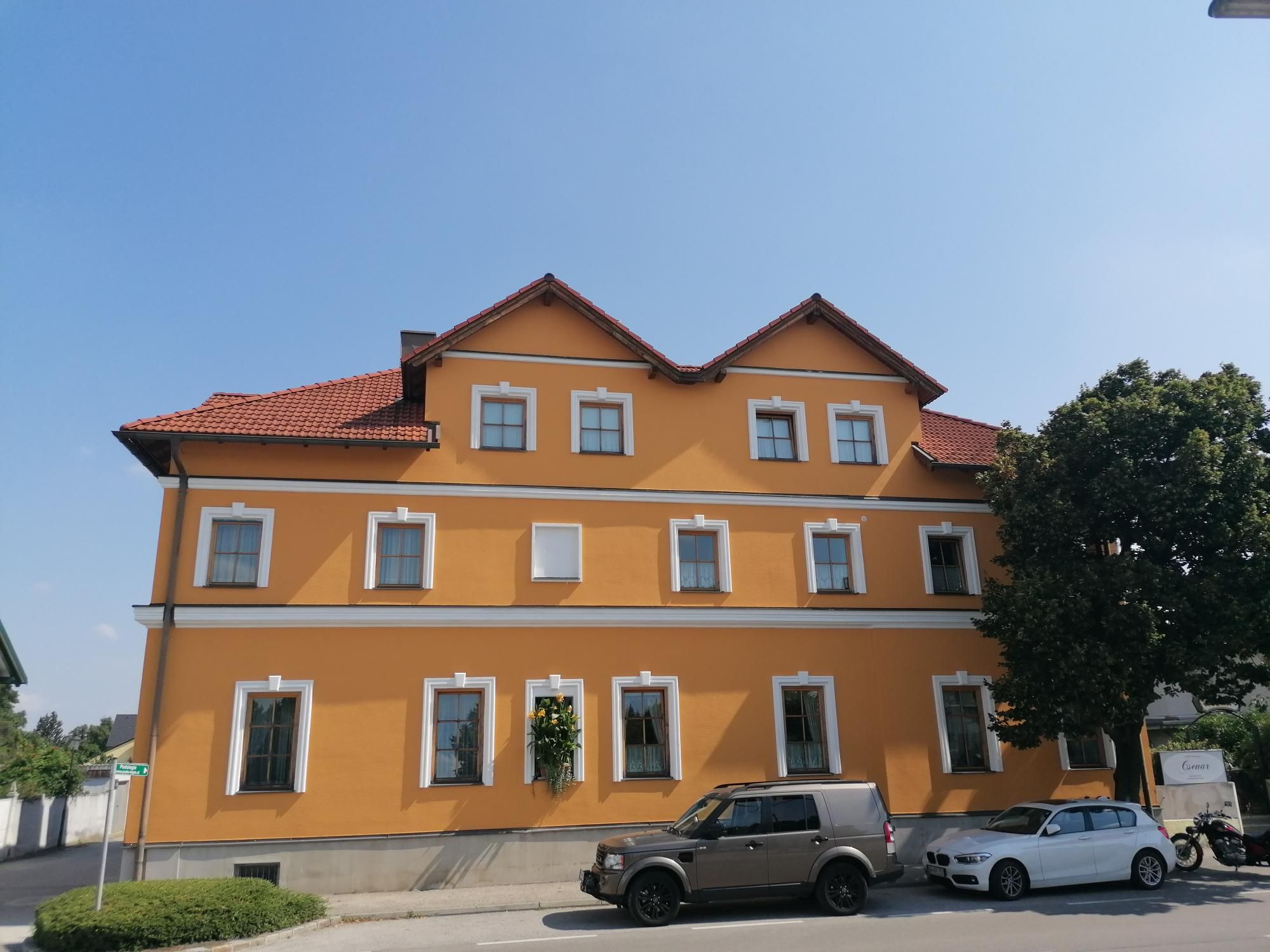 Žlutá budova s červenými střešními taškami a několika okny, před ní zaparkovaná auta a strom.