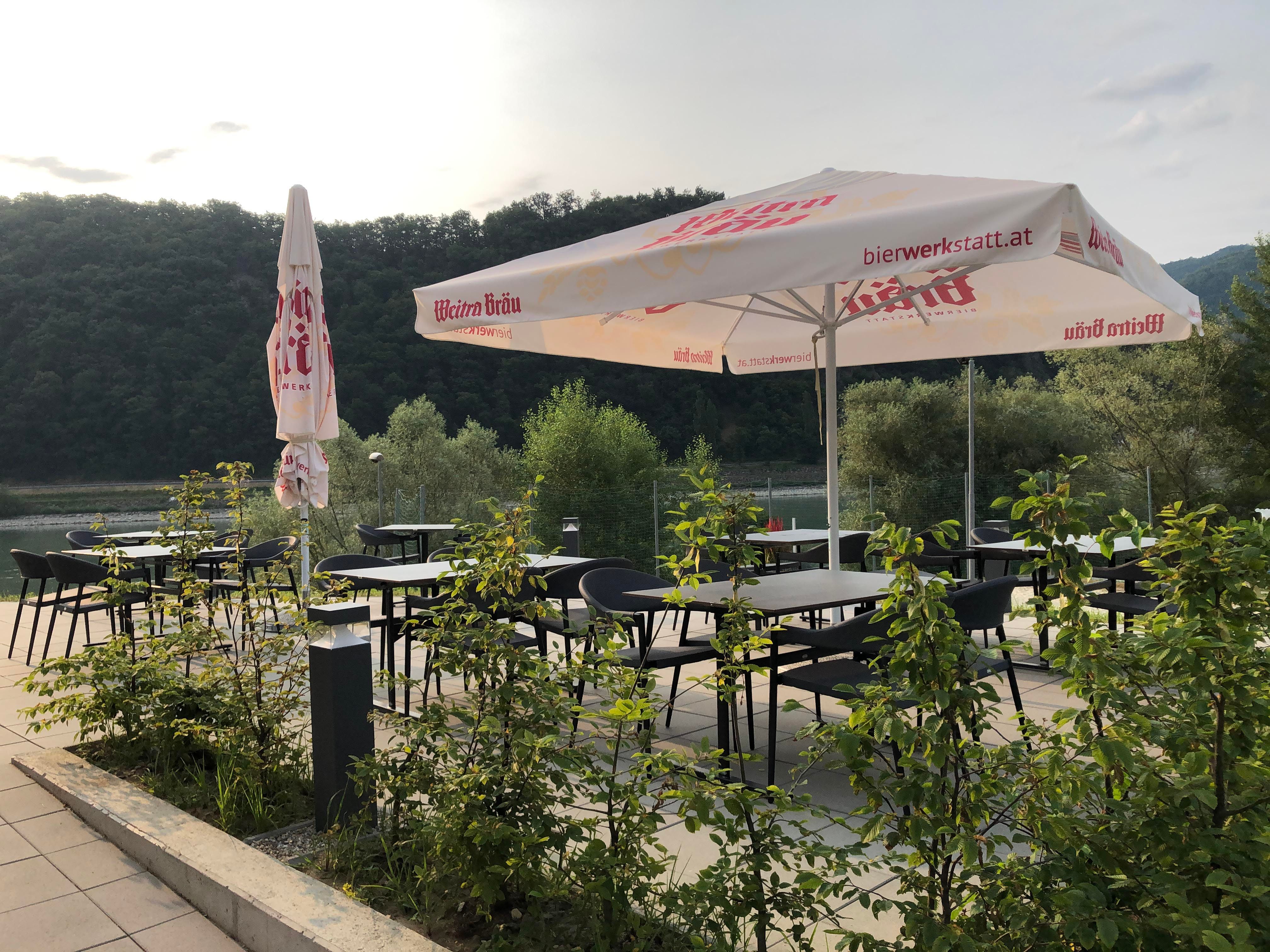 Terasa se stoly, židlemi a slunečníky, obklopená přírodou.