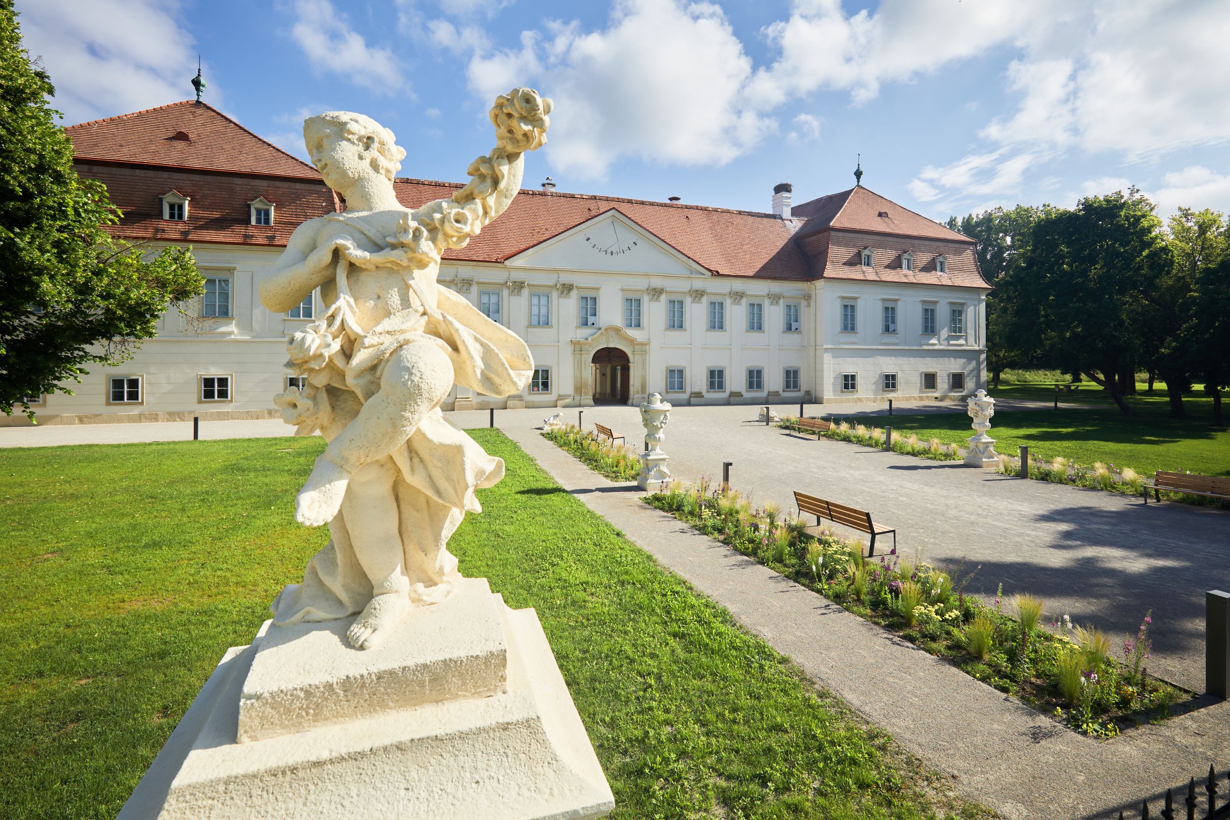 Barokní socha před historickou budovou s červenou střechou a udržovanou zahradou.