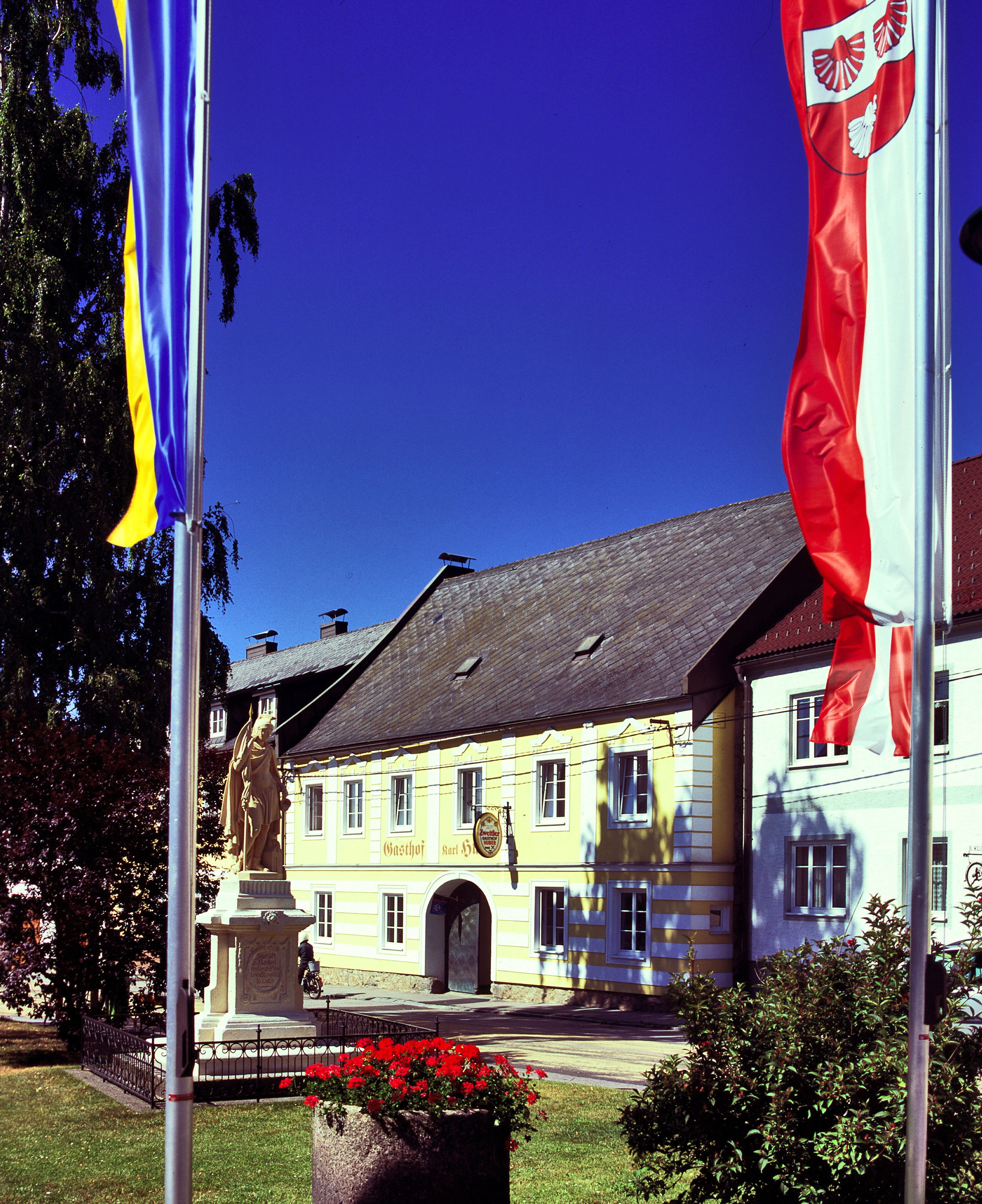 Žlutá budova s nápisem "Gasthof Karl Huber", obklopená vlajkami a sochou.
