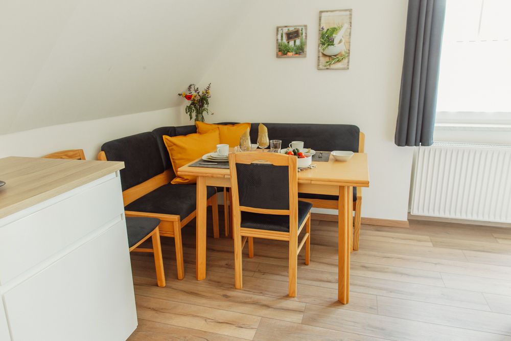 Jídelní kout s dřevěným stolem, židlemi a rohovou lavicí, prostřeným nádobím, kyticí květin a dekorací na stěně.