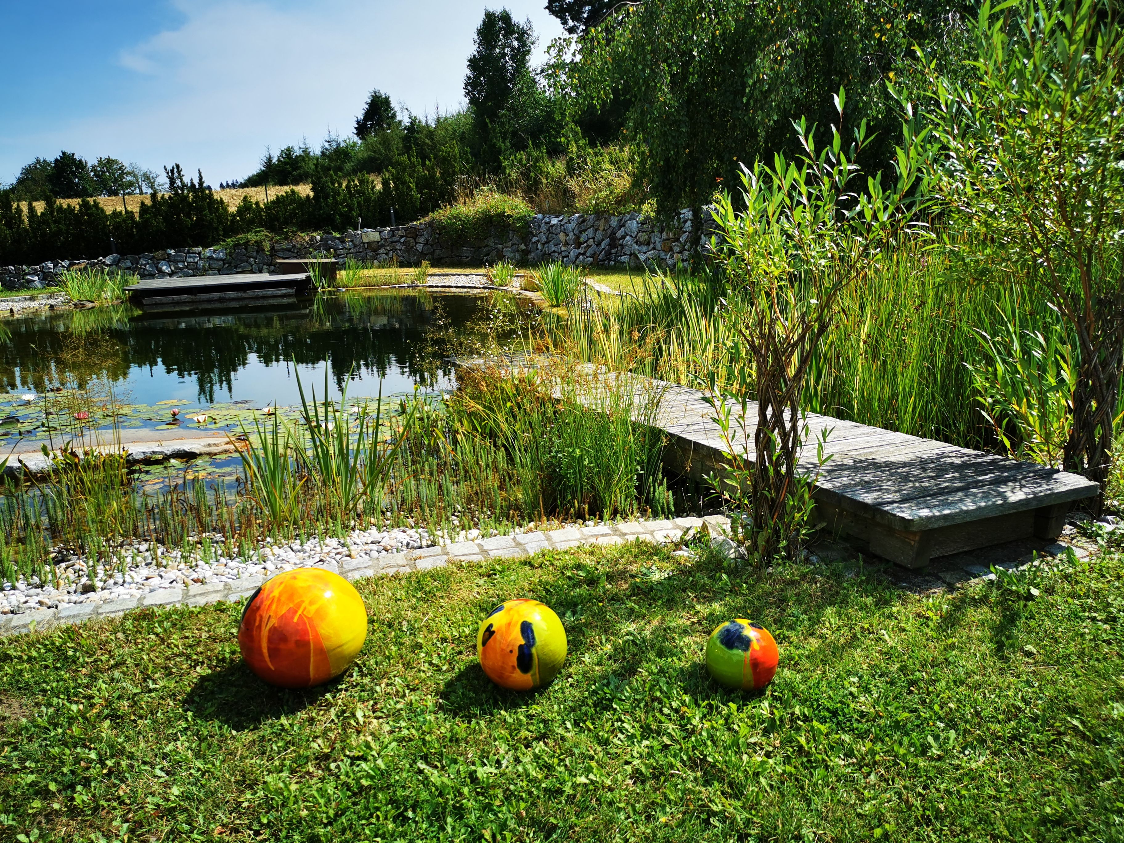 Přírodní koupací jezírko s dřevěným chodníkem a barevnými míči na trávníku.