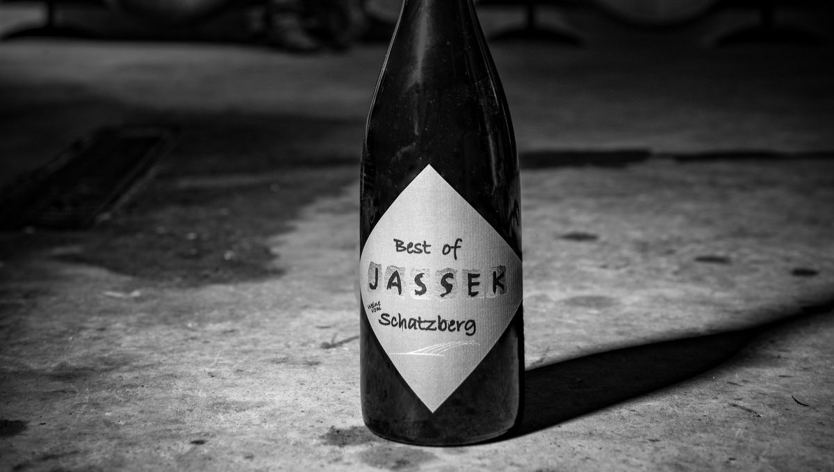 Černobílá fotografie láhve vína s nápisem "Best of Jassek Schatzberg" na betonové podlaze.