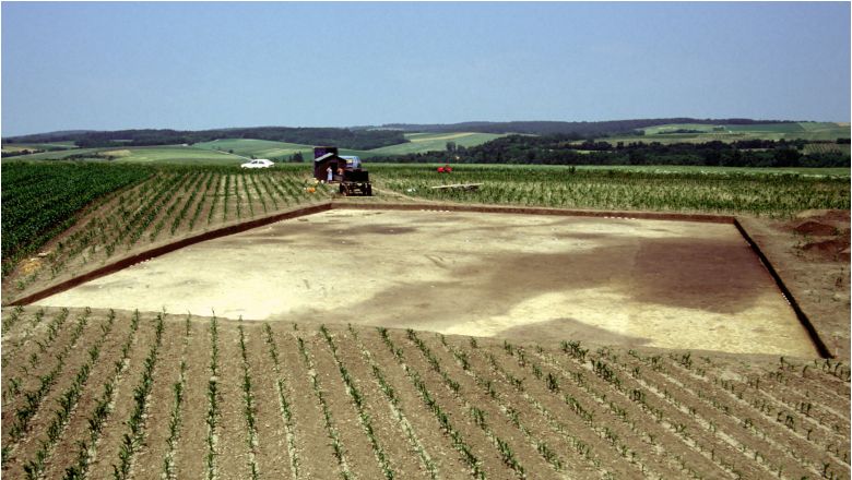 Archeologické vykopávky na poli se zemědělskými stroji v pozadí.