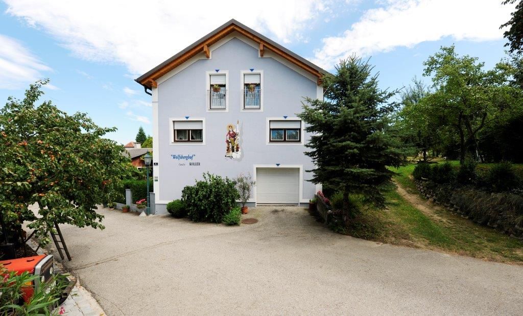 Dvoupatrový dům s modrou fasádou a dřevěnou střechou, obklopený stromy a dlážděnou cestou.