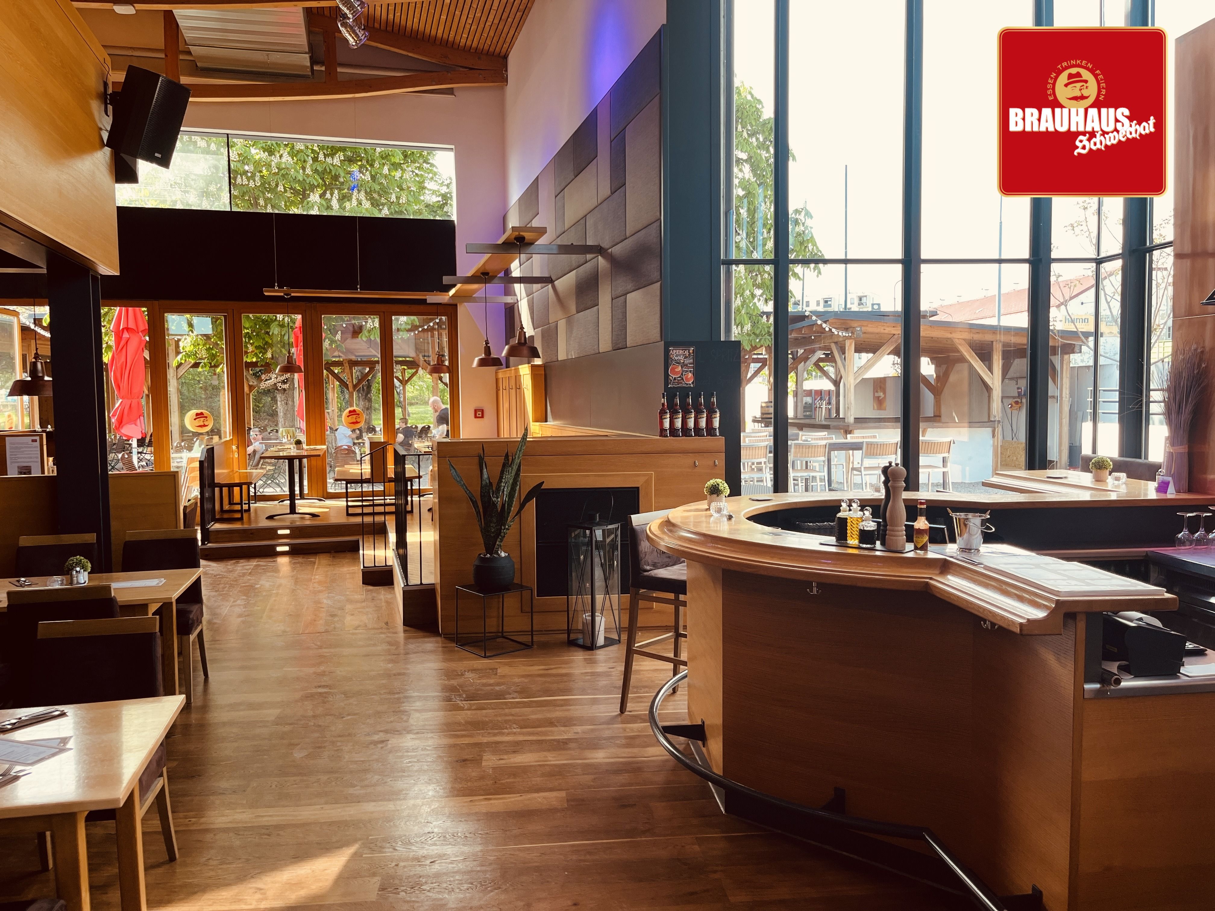 Pohled do interiéru moderního pivovaru s dřevěným nábytkem a velkými okny.