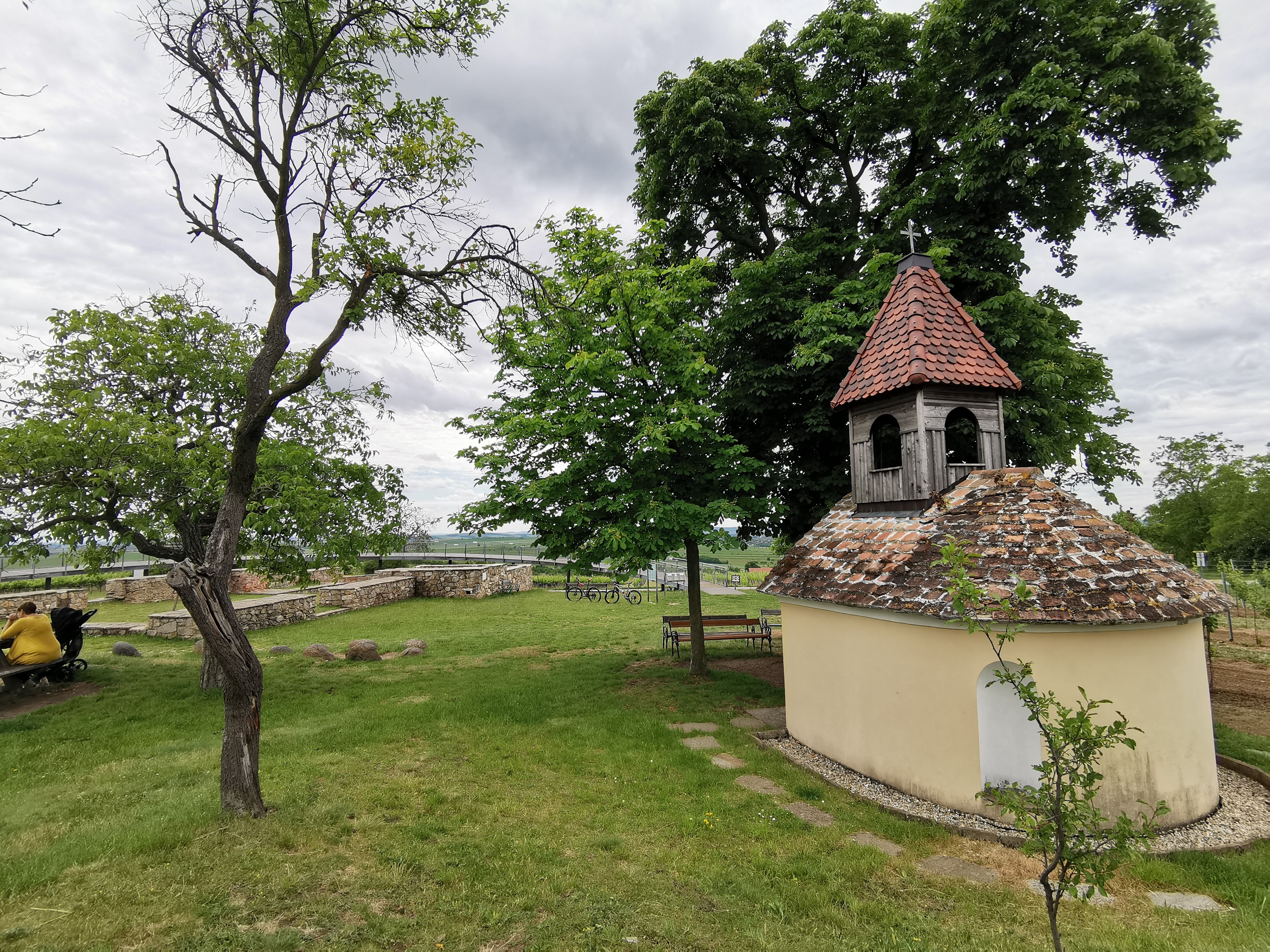 Malá kaple s červenou taškovou střechou v zelené krajině, obklopená stromy a lavičkami.