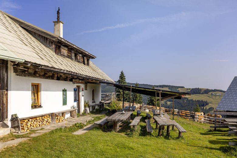 Tradiční horská chata s dřevěnými stoly a lavicemi venku, obklopená zelenými loukami a zalesněnými kopci pod modrou oblohou.