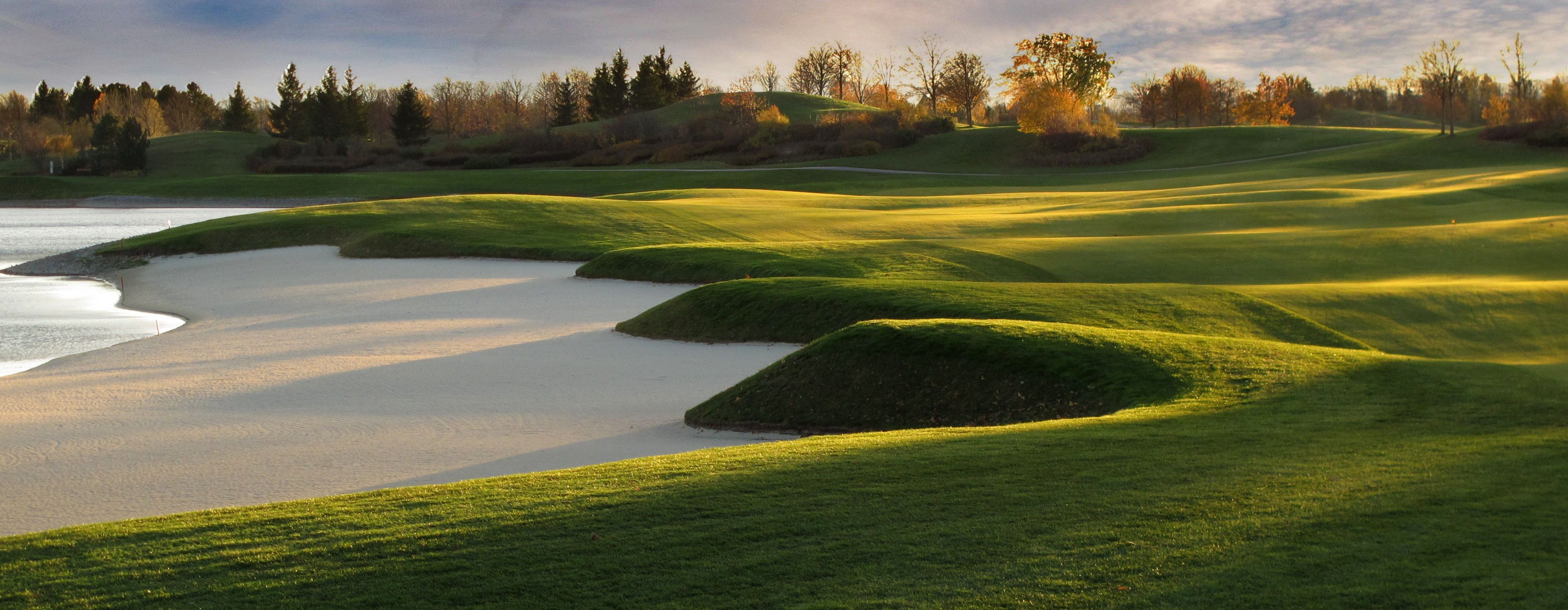 Panorama golfového hřiště s písečnými bunkry a zelenými kopci ve slunečním světle.