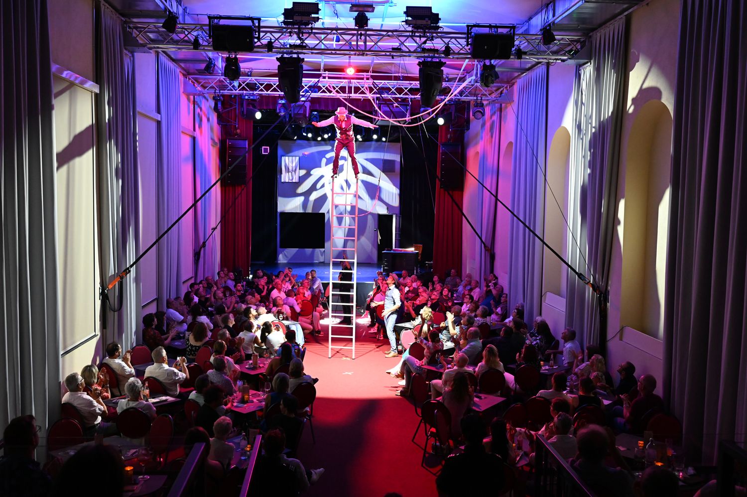 Umělec balancuje na žebříku v přeplněném divadle s barevným osvětlením.