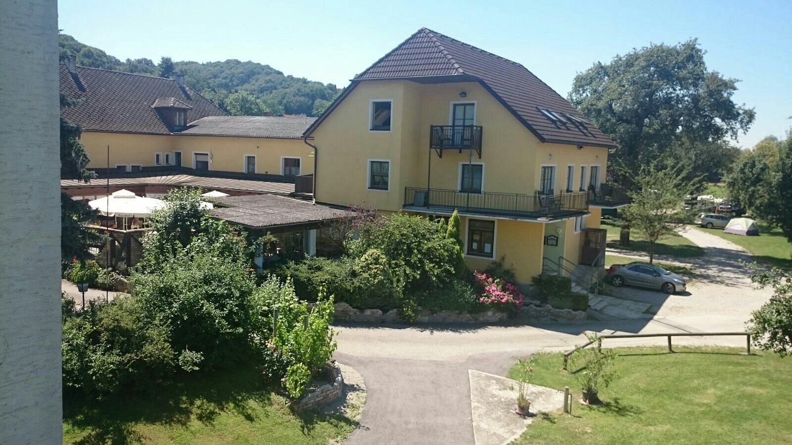 Žlutá budova s terasou a zahradou, obklopená stromy a autem na parkovišti.