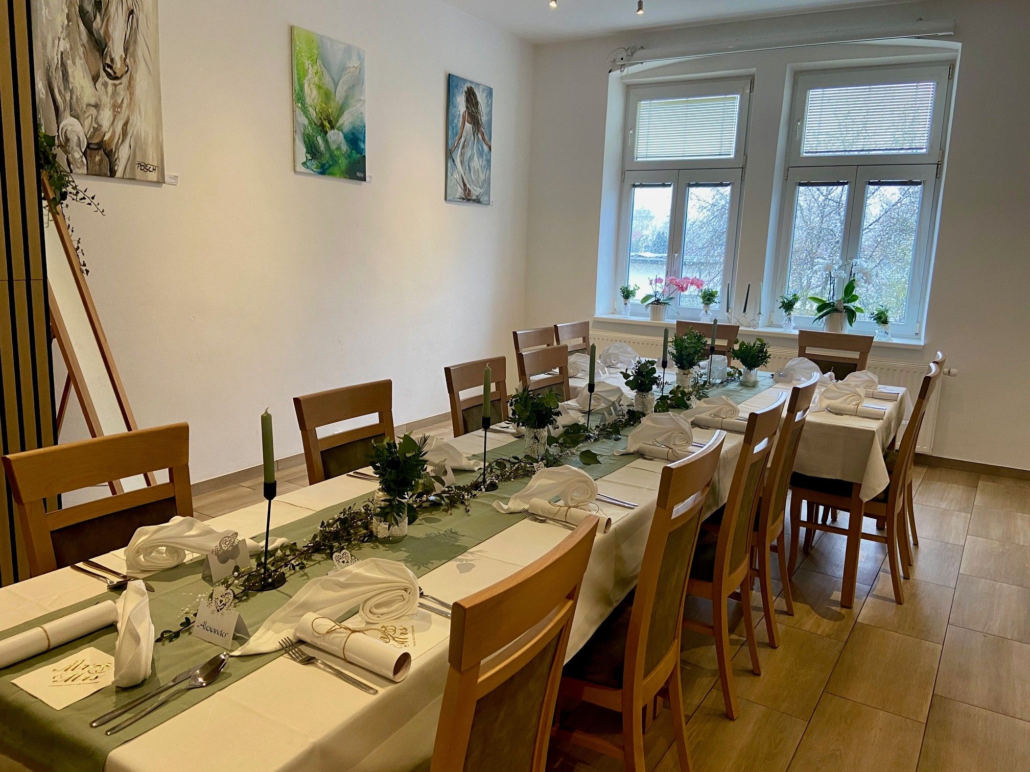 Slavnostně prostřený stůl v místnosti s uměleckými díly na stěnách a velkými okny.