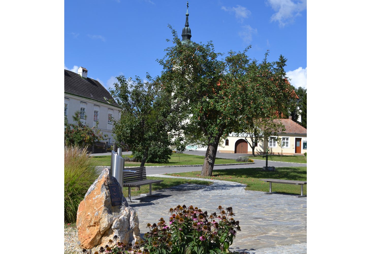 Vesnická náves v Hagenbergu s kostelem, stromy a lavičkami.