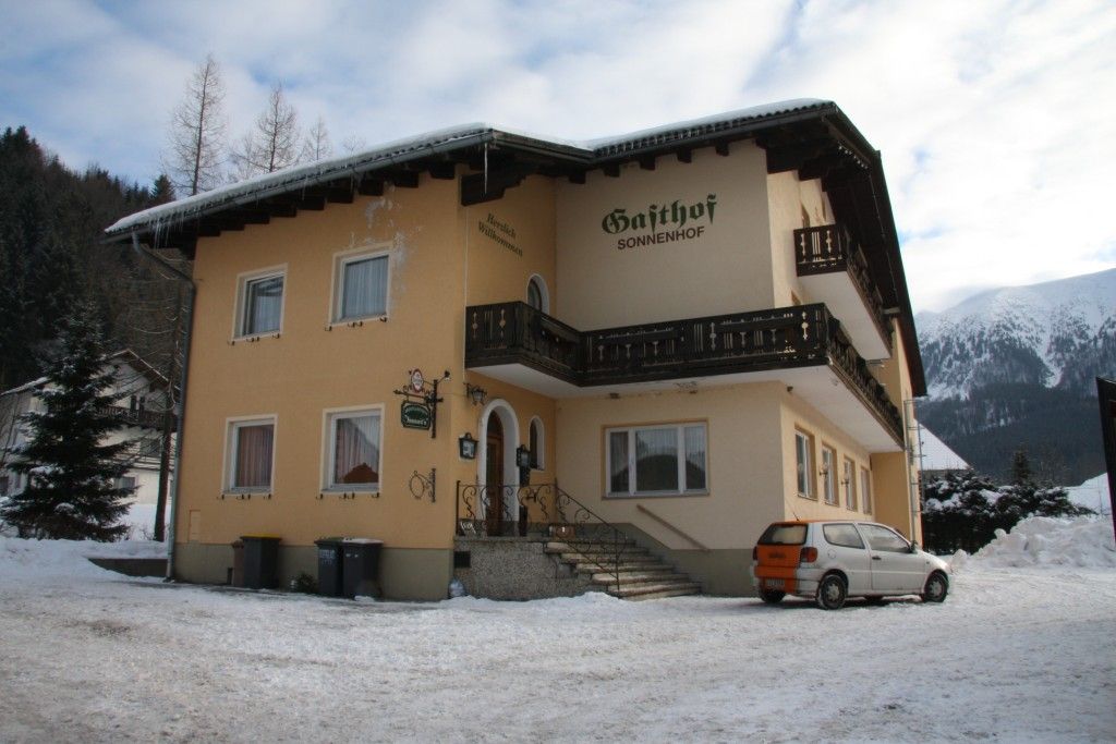 Žlutá budova s nápisem "Gasthof Sonnenhof" na sněhu, obklopená horami.