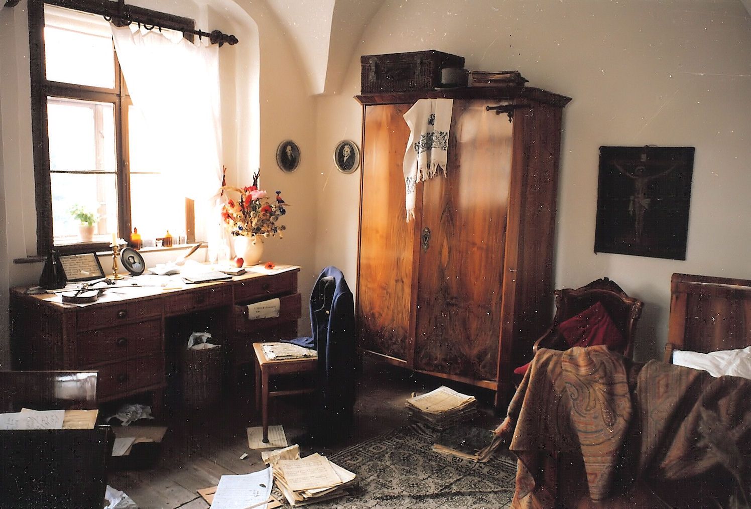 Historický pokoj s dřevěným stolem, notami, skříní a postelí. Pokoj působí neuspořádaně a staromódně.