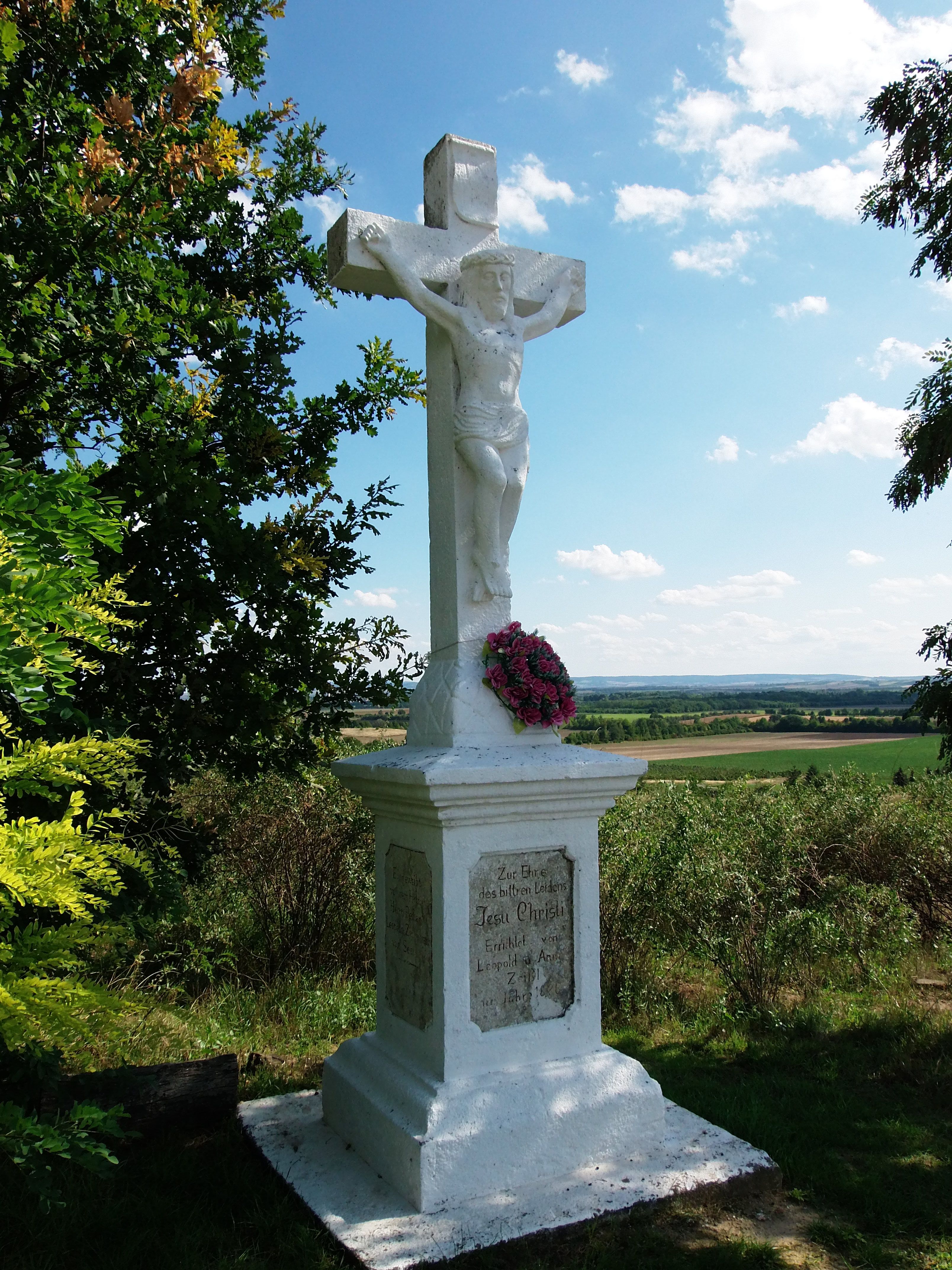Bílý kamenný kříž s postavou Ježíše a květinovým věncem, obklopený stromy a krajinou v pozadí.