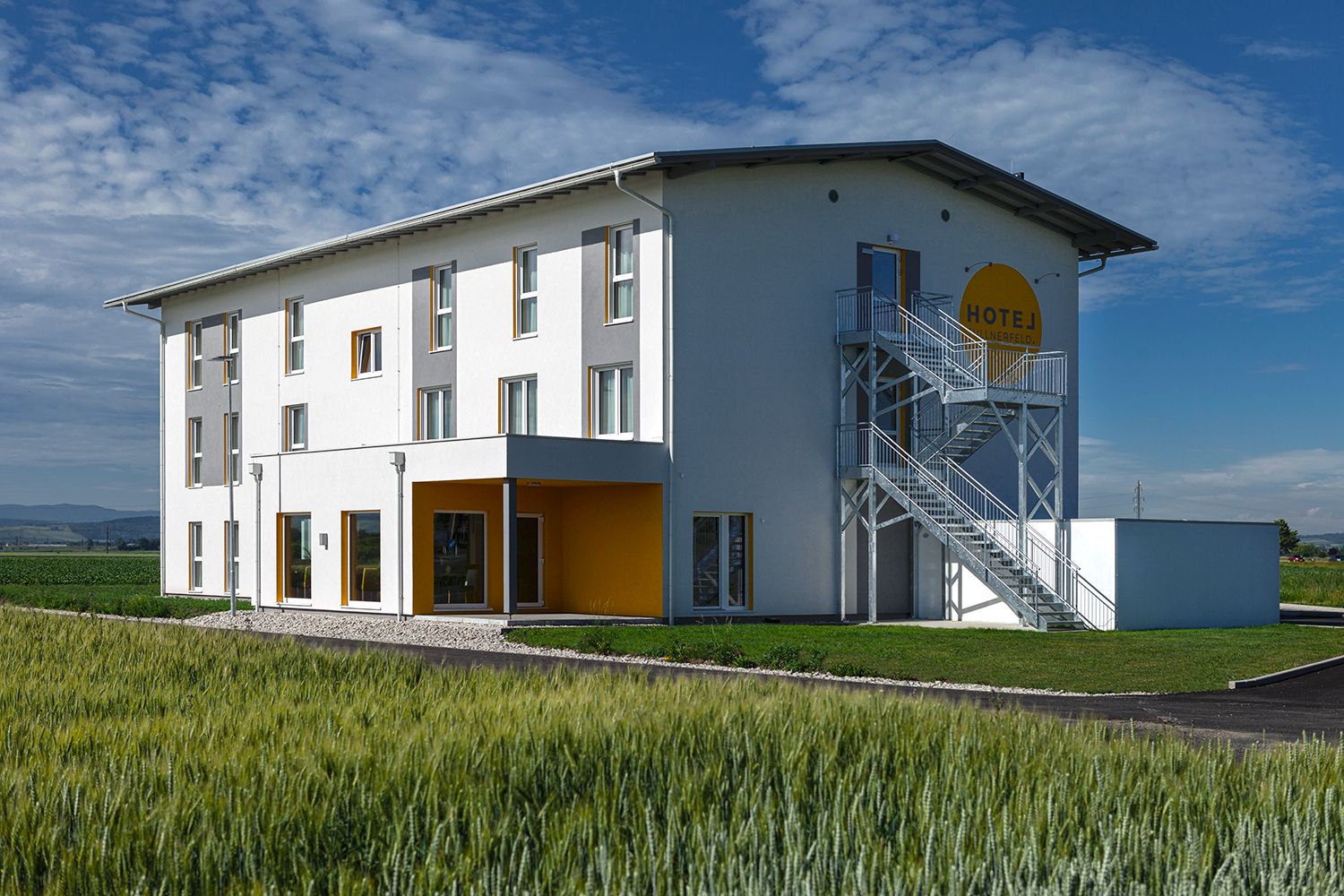 Pohled zvenčí na moderní hotel s bílou fasádou a žlutými akcenty, obklopený poli.