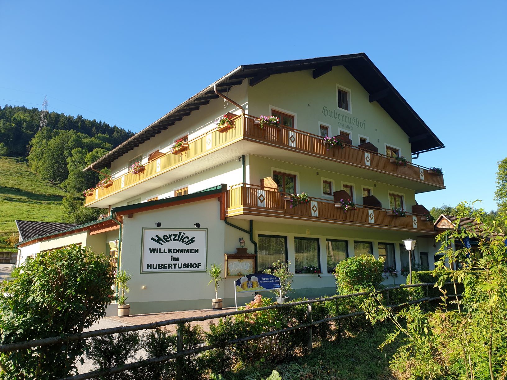 Tradiční hostinec Hubertushof s květinami na balkonech, obklopený zelenou krajinou a modrou oblohou.