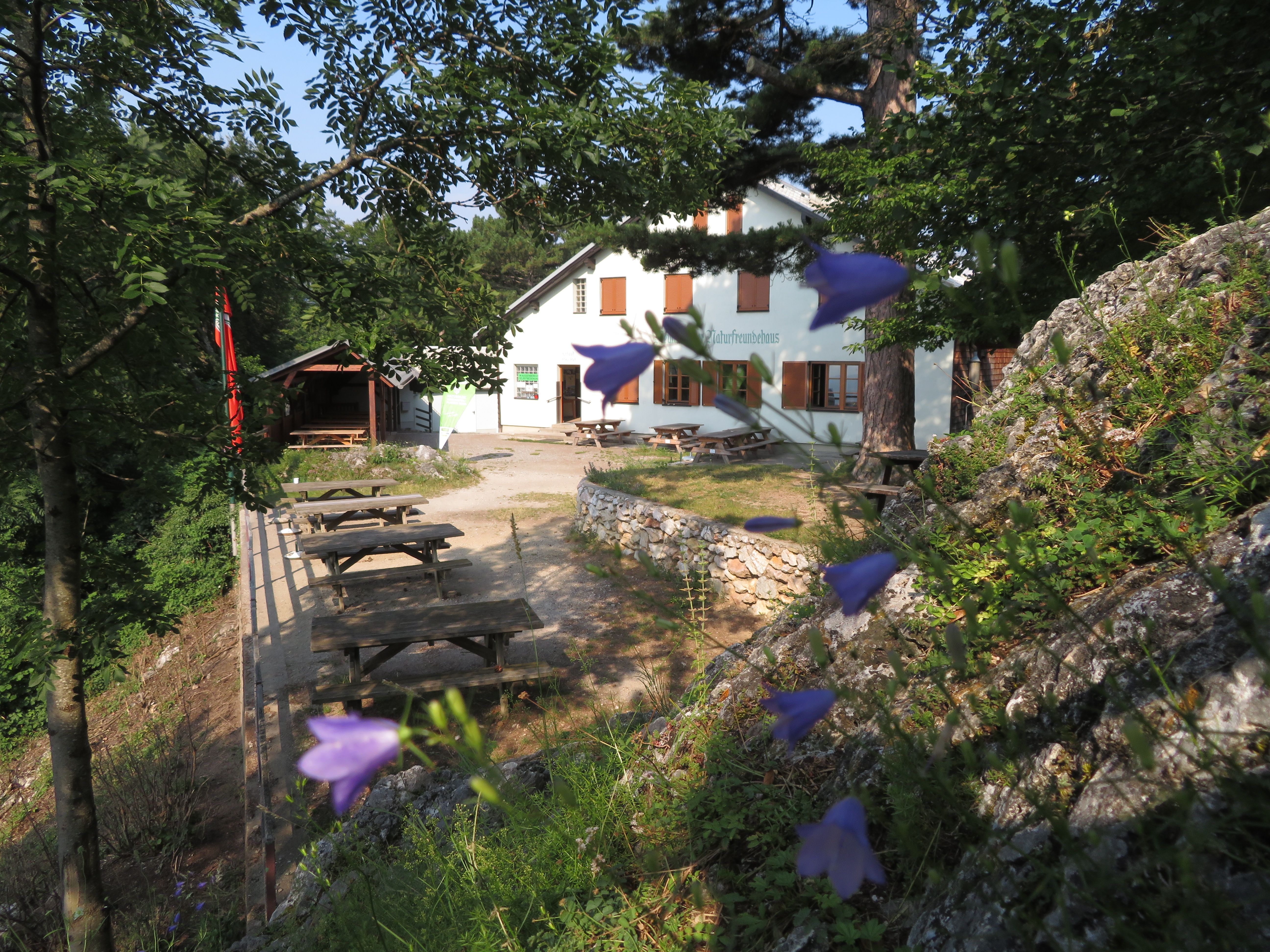 Bílá budova s nápisem "Naturfreundehaus" obklopená stromy a květinami. V popředí jsou vidět dřevěné lavice a stoly.