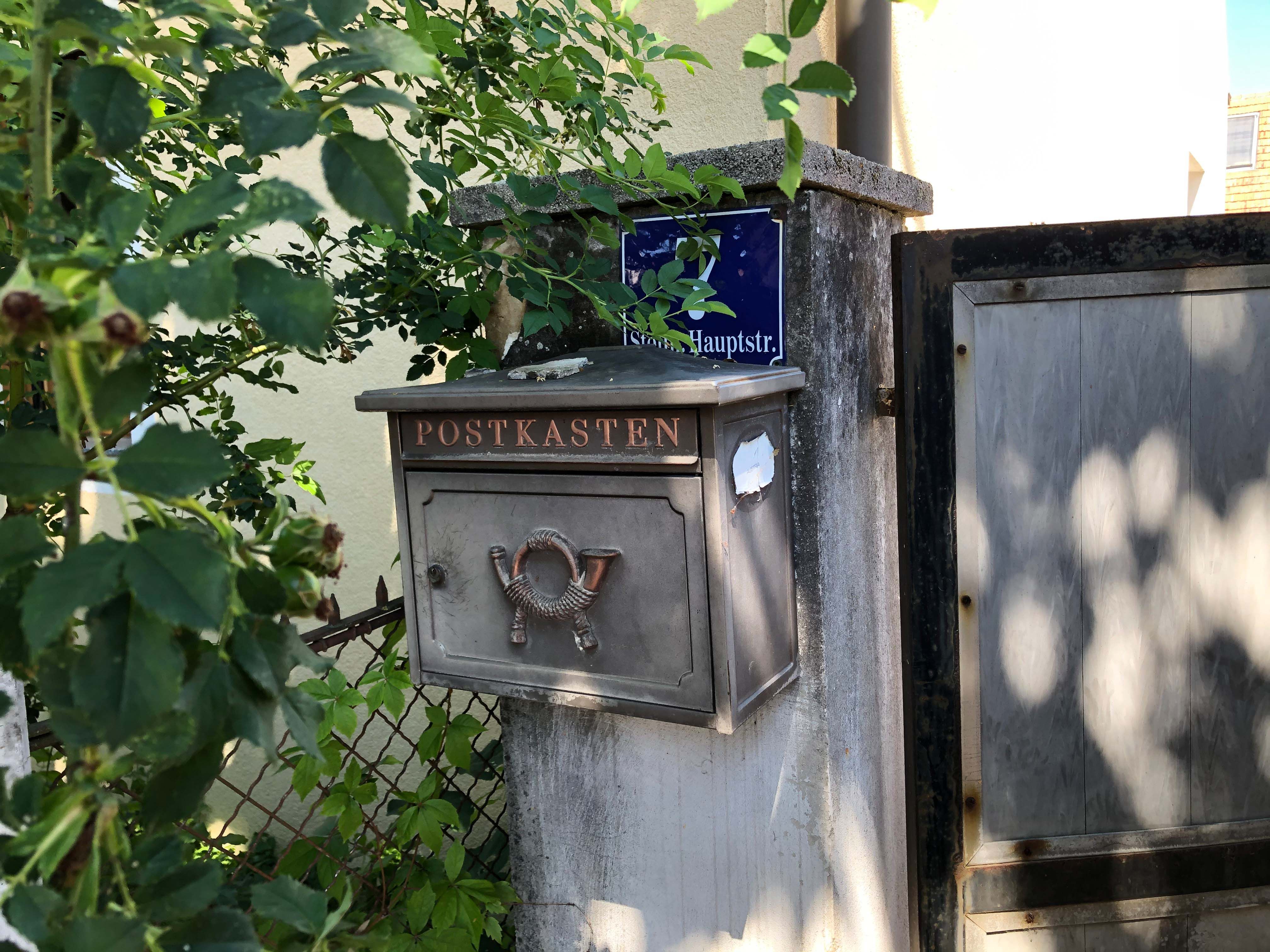 Stará poštovní schránka s nápisem "Postbox" na zdi vedle brány, obklopená rostlinami.