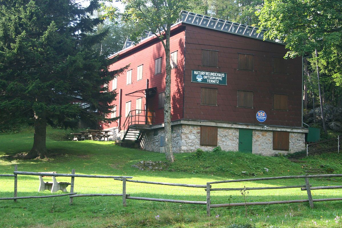 Červená budova s dřevěným obložením, obklopená stromy a loukou, s nápisem "Naturfreundehaus Ortsgruppe Ternitz".