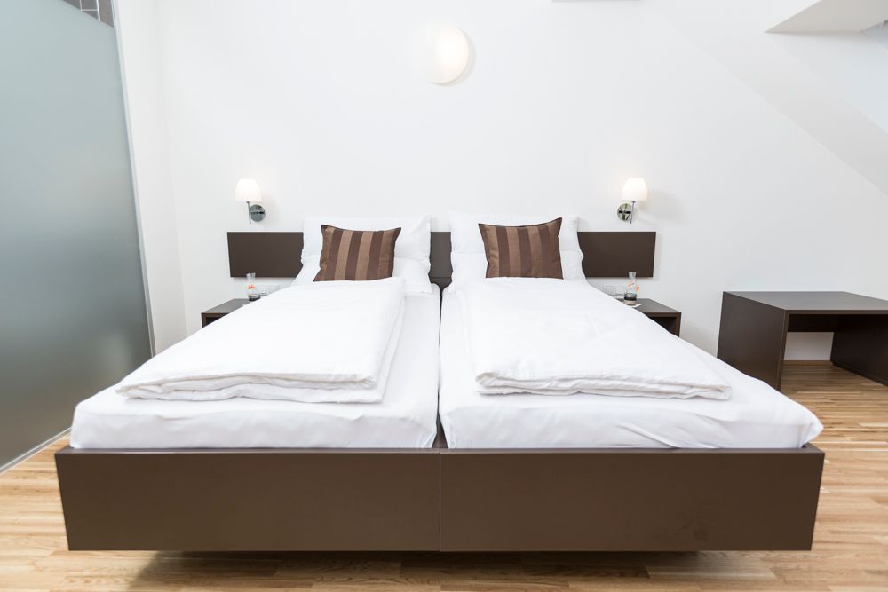 Moderní pokoj pro hosty se dvěma samostatnými postelemi, bílými peřinami a hnědými polštáři, dřevěnou podlahou a nočními stolky s lampičkami.