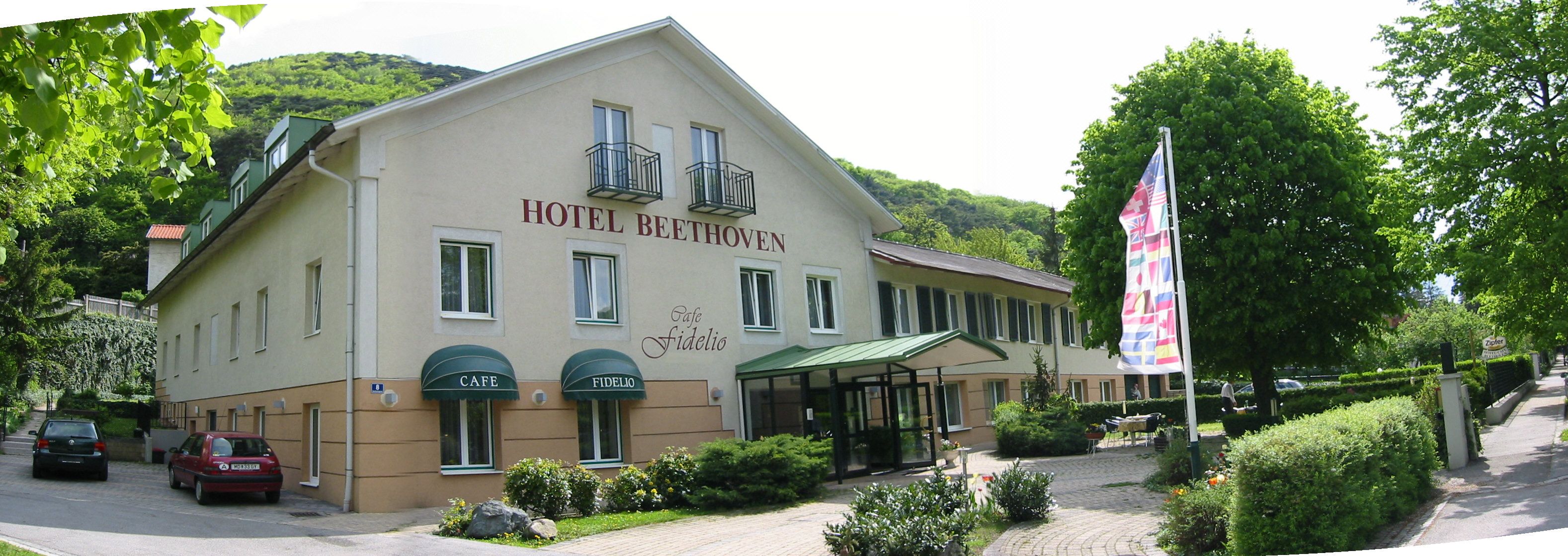 Pohled na exteriér hotelu Beethoven s kavárnou Fidelio, obklopenou stromy a vlajkami.