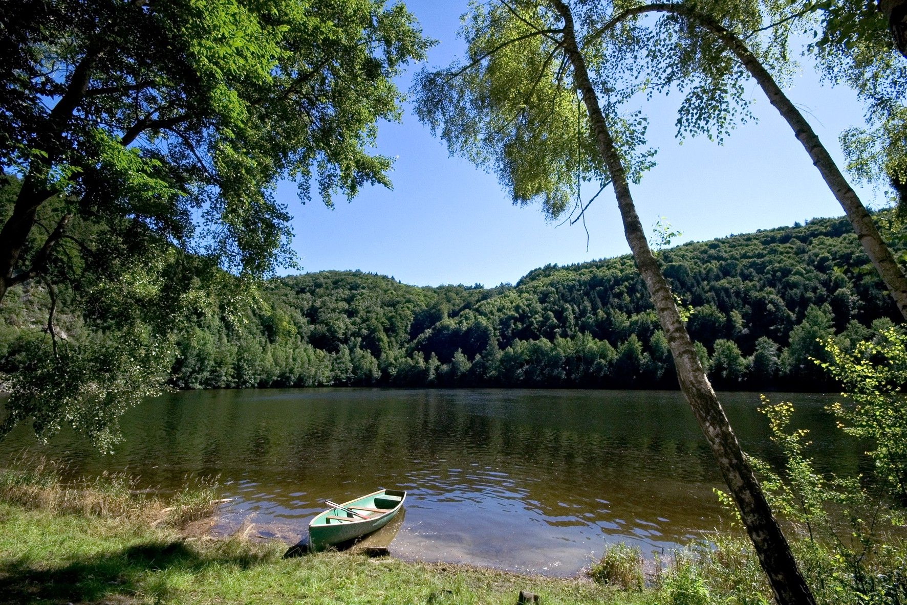 Klidné jezero s veslicí na břehu, obklopené hustě zalesněnými kopci a jasnou oblohou.