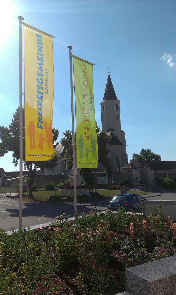 Dvě barevné vlajky s nápisy "Freizeitgemeinde Langau" a "Do bin i daham" před kostelem.