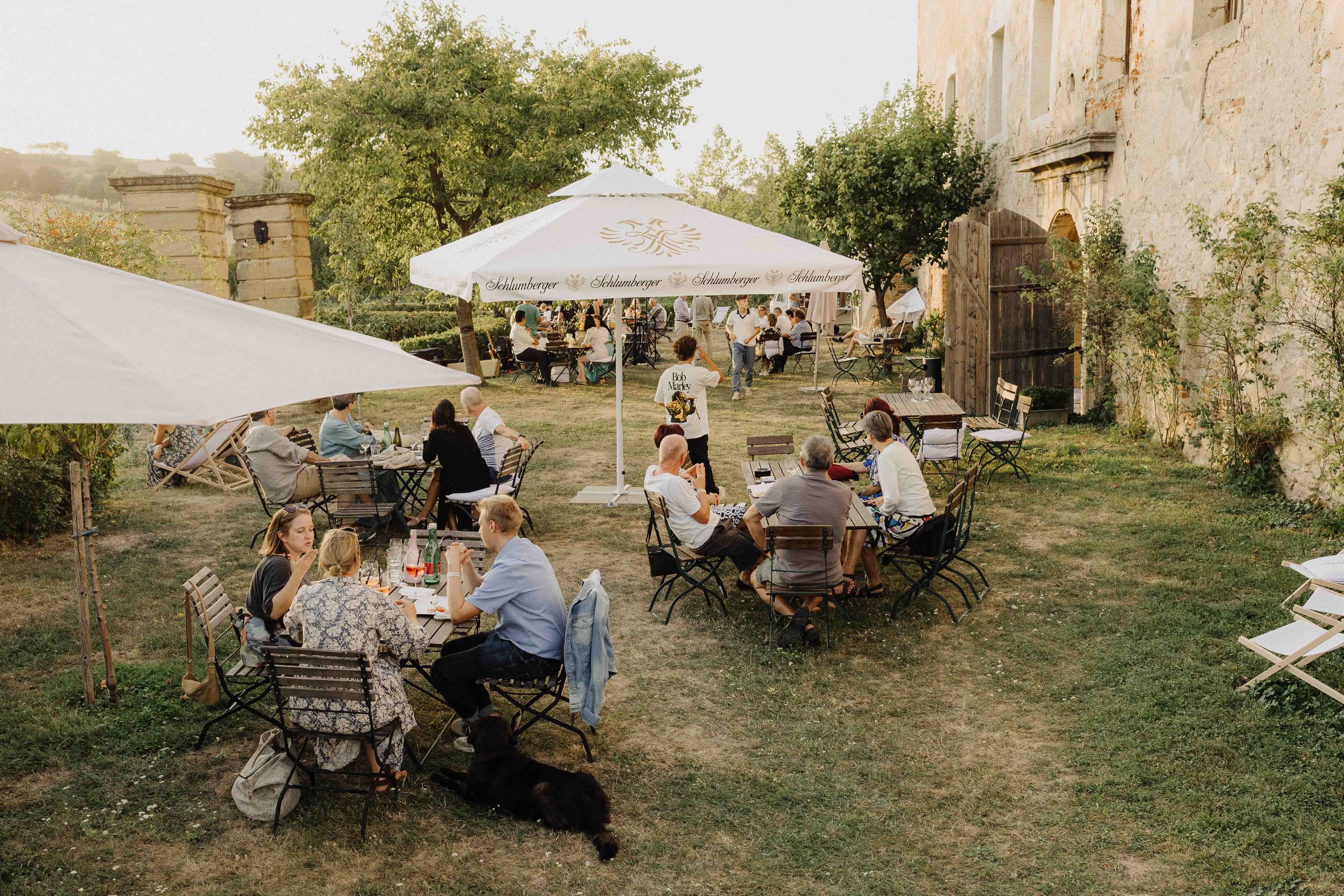 Lidé sedí u venkovních stolů na zahradě se slunečníky.