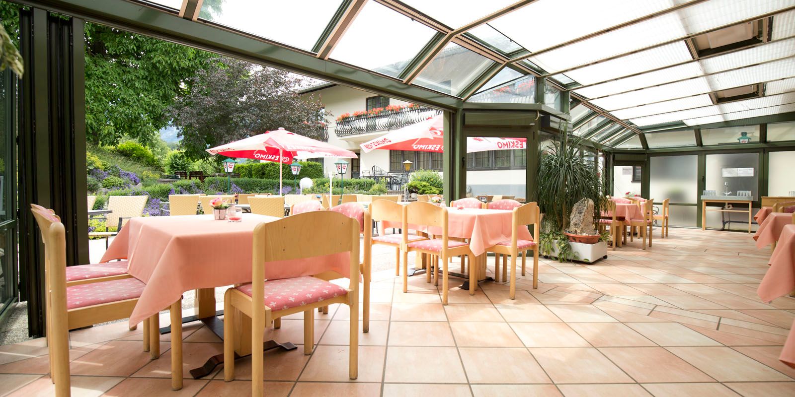 Světlá restaurace s prosklenou střechou, růžovými ubrusy a výhledem do zahrady se slunečníky.