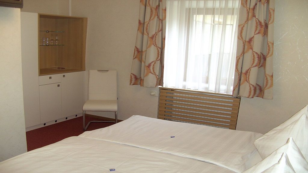 Hotelový pokoj s postelí, židlí, skříní a oknem se závěsy.