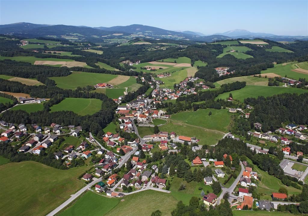Letecký pohled na Zöbern, venkovskou obec s domy a poli, obklopenou kopci a lesy.