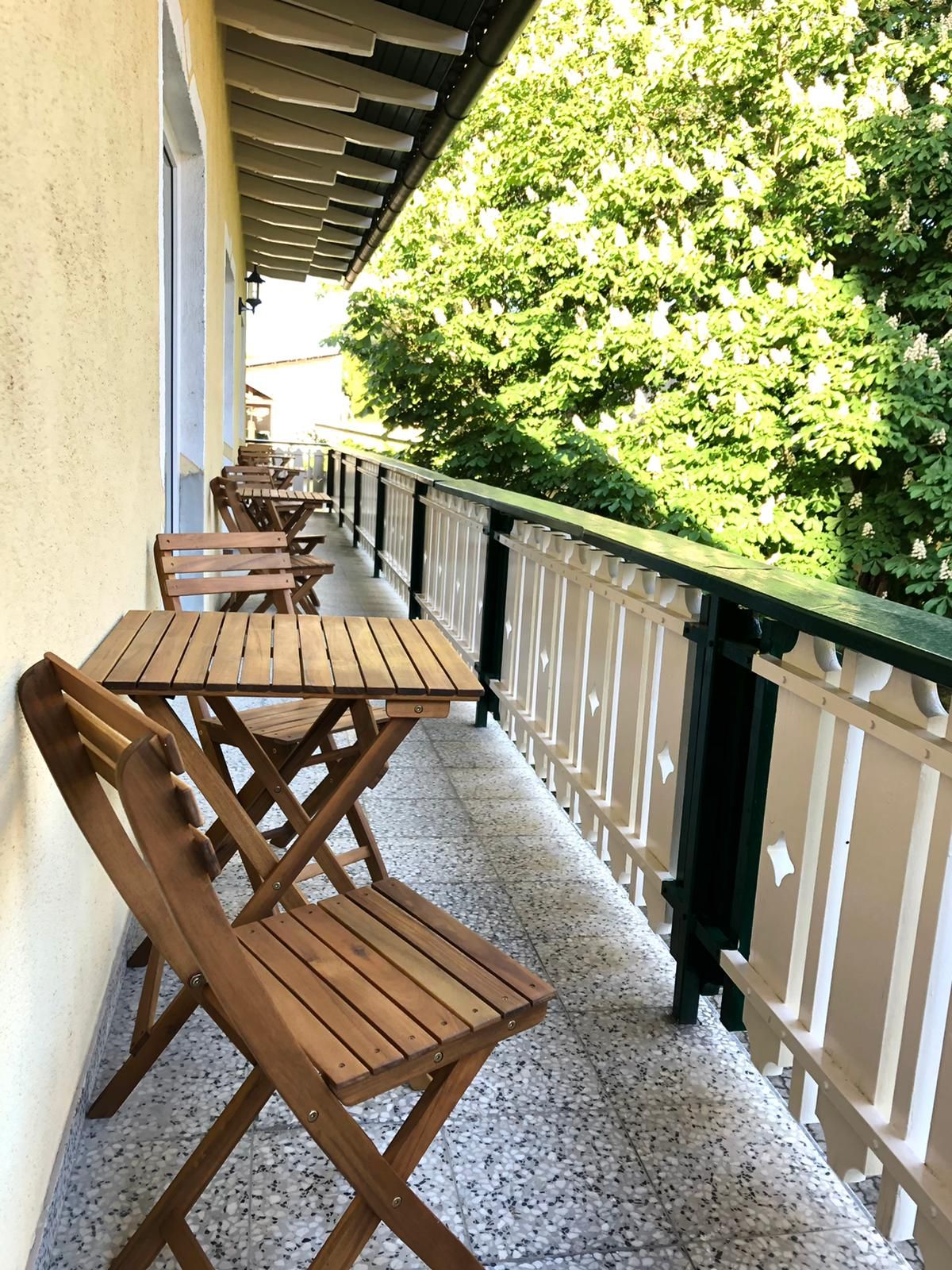 Dřevěný balkon se stolem a židlemi, vedle něj zelené stromy.
