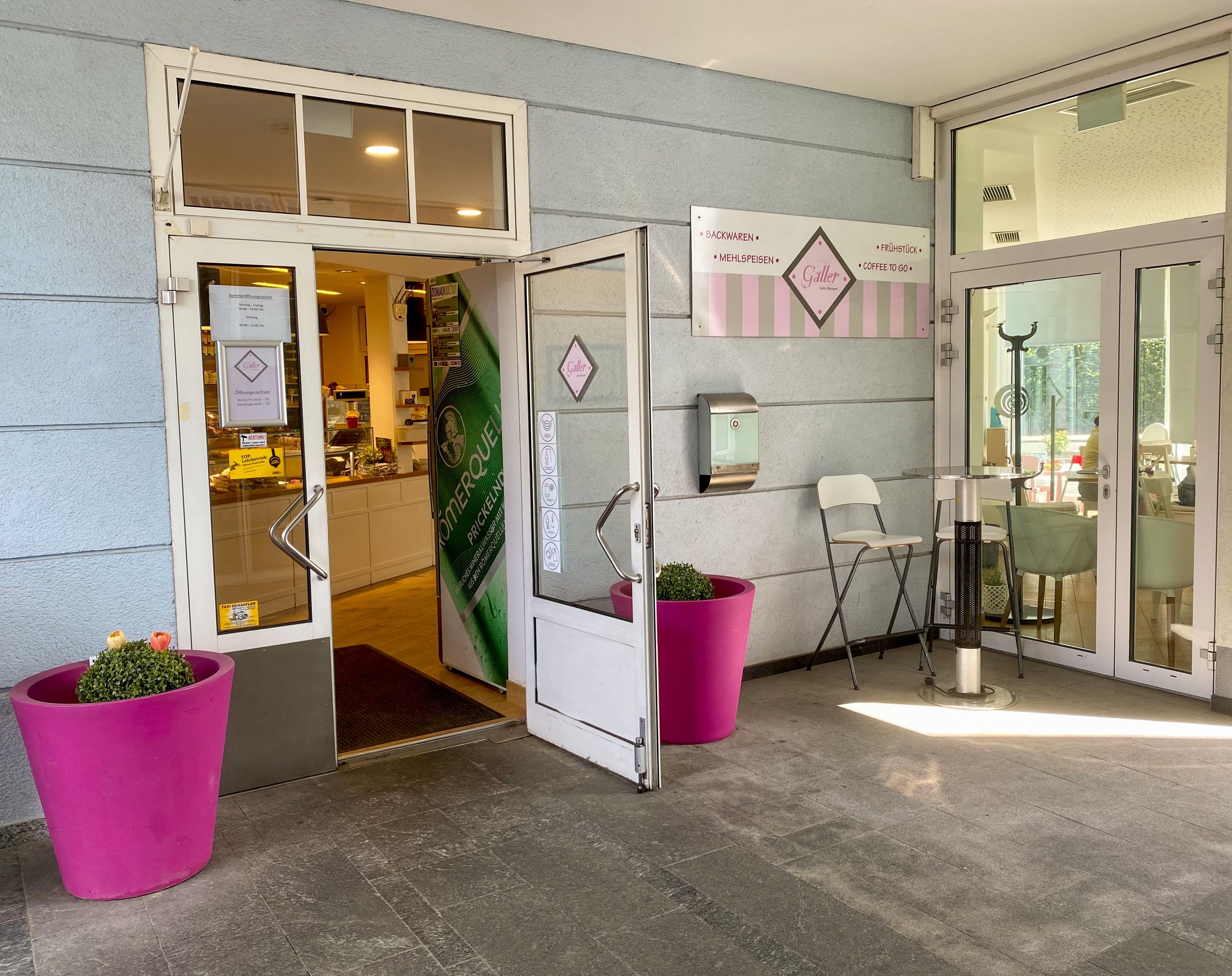 Vchod do kavárny a vpravo růžové květináče před světle modrým domem 