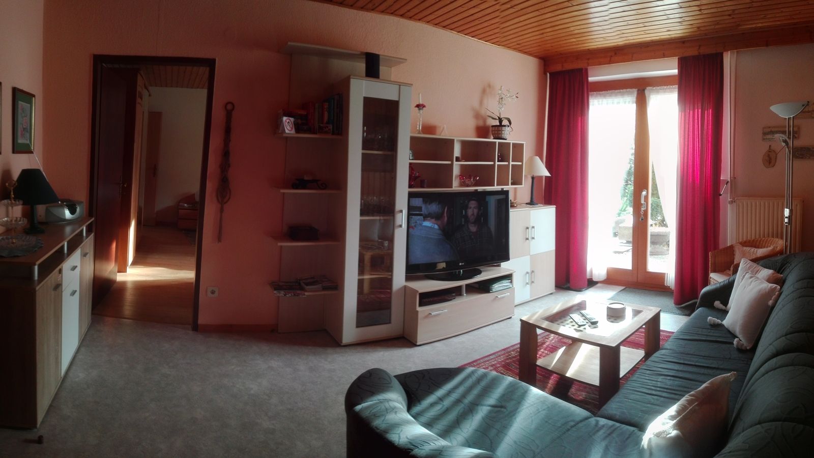 Útulný obývací pokoj s pohovkou, TV, poličkou a skleněnými dveřmi s červenými závěsy.