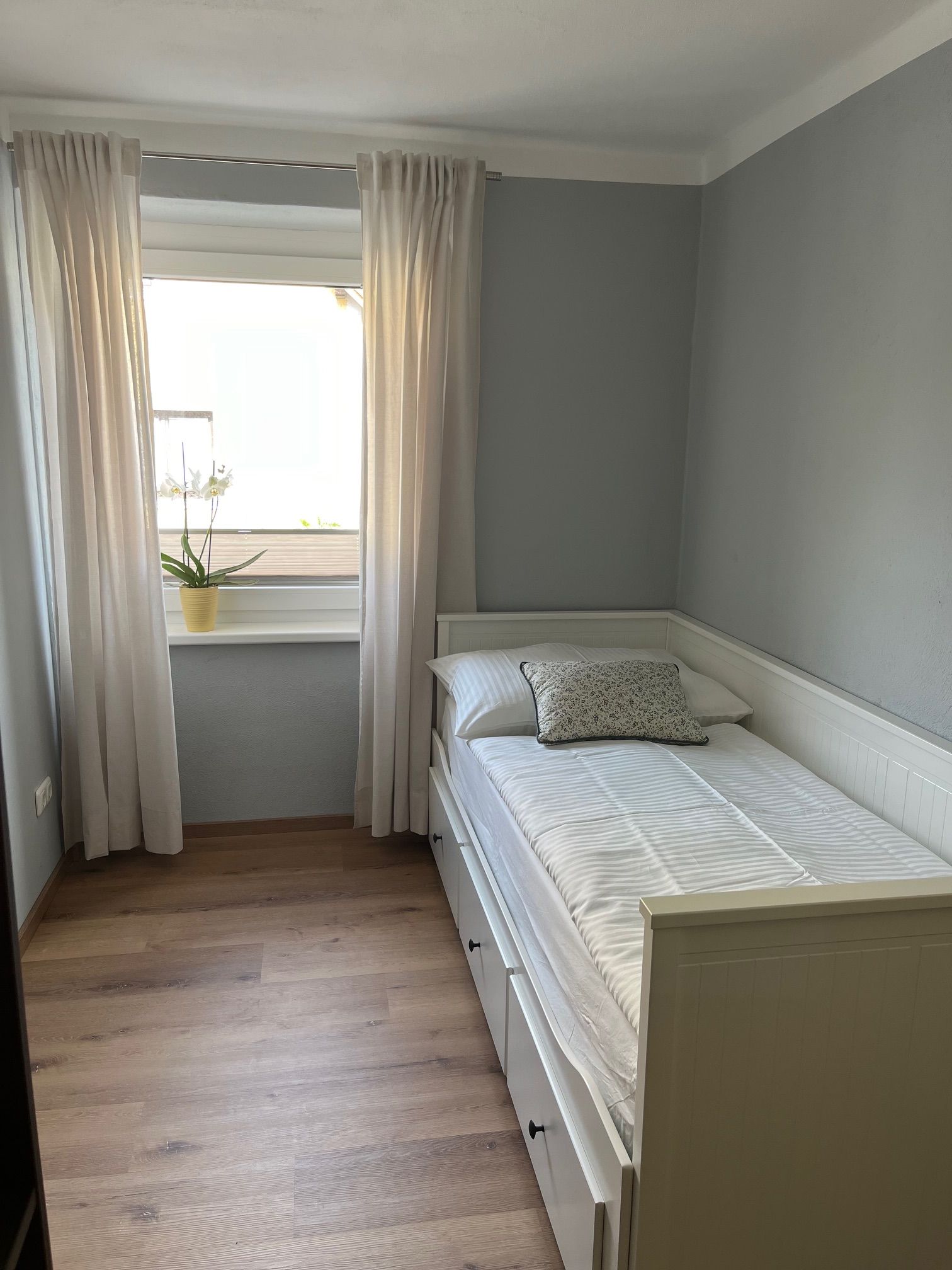 Malá ložnice s jednolůžkem, bílými závěsy a rostlinou na parapetu.