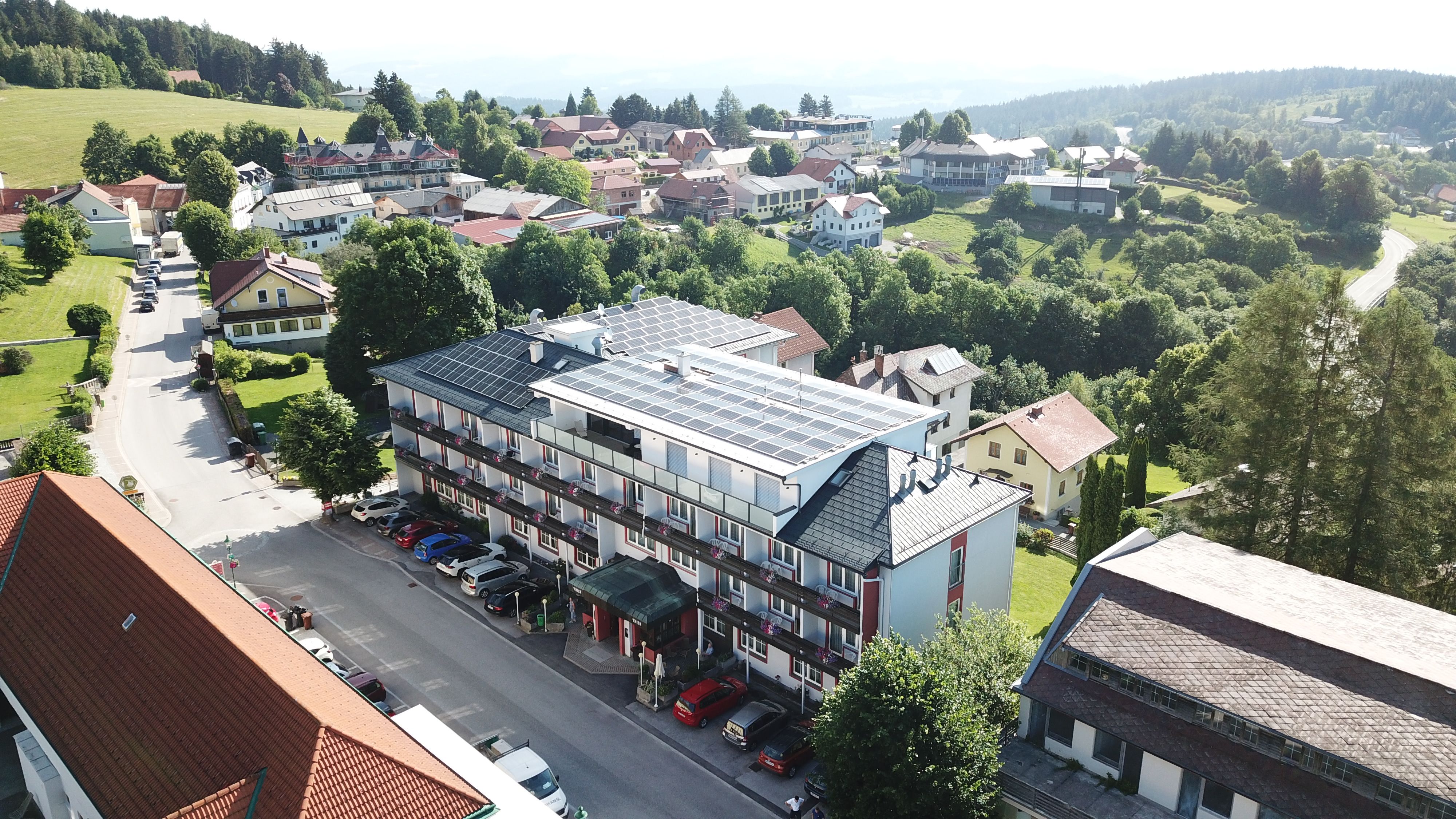 Letecký snímek hotelu se solárními panely na střeše, obklopeného stromy a dalšími budovami v venkovské oblasti.