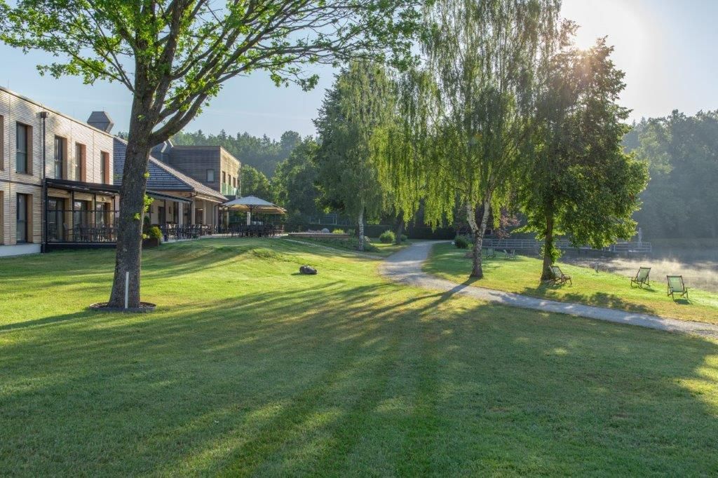 Zelená louka se stromy, budovami a lehátky na břehu jezera v záři slunce.