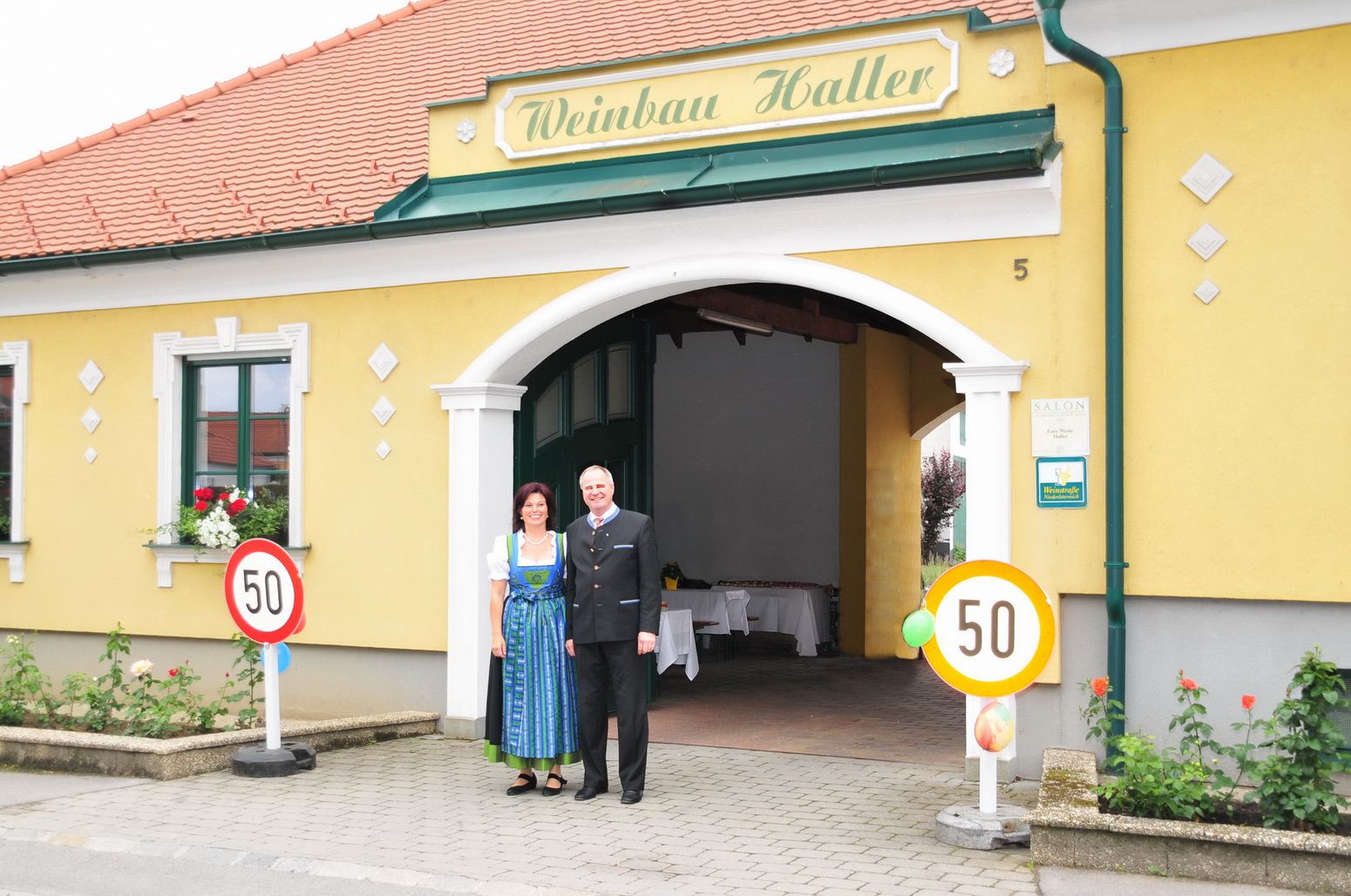 Před žlutou budovou s nápisem "Weinbau Haller" stojí manželský pár.
