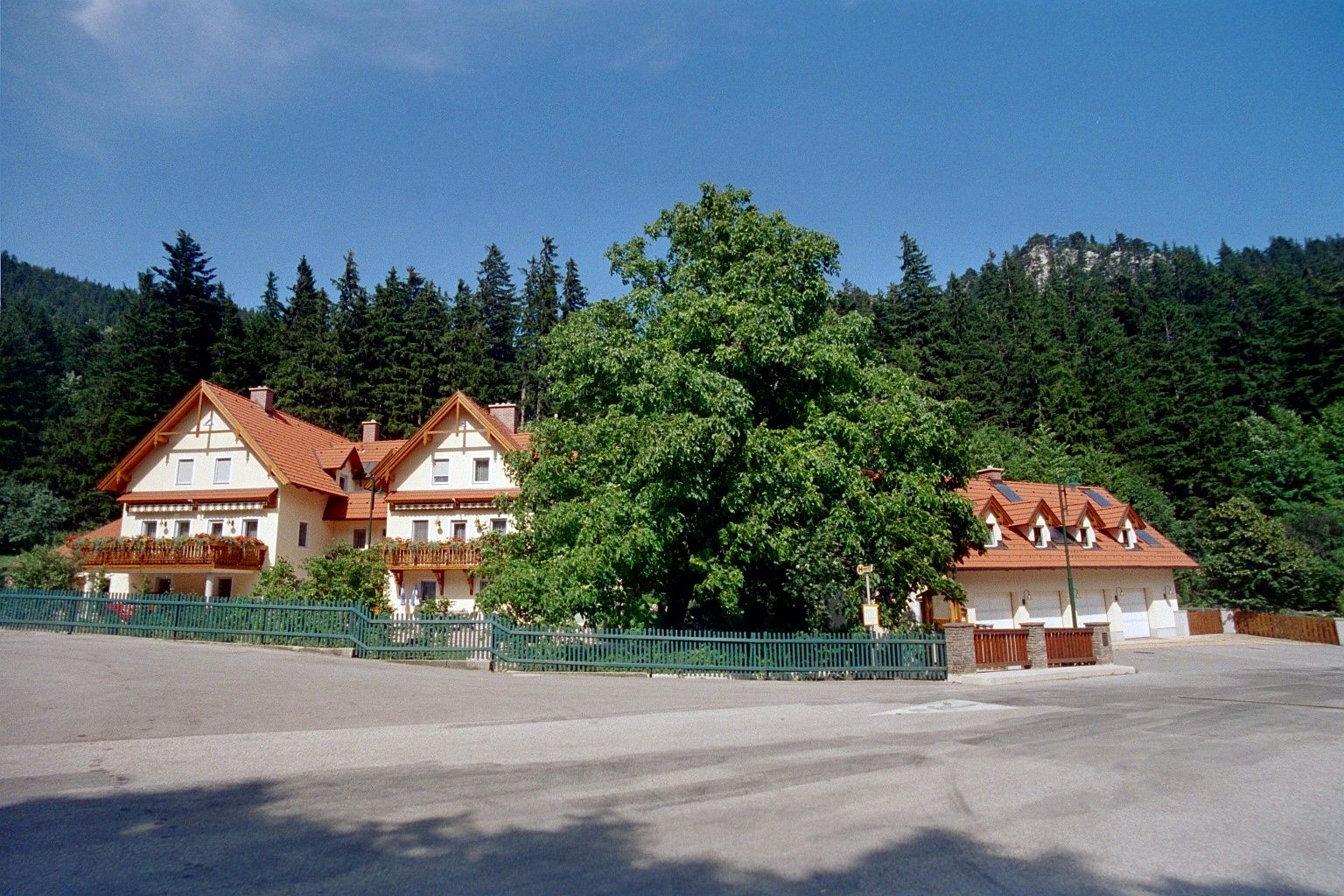 Velký dům s červenými střechami a balkony, obklopený stromy a lesem v pozadí.