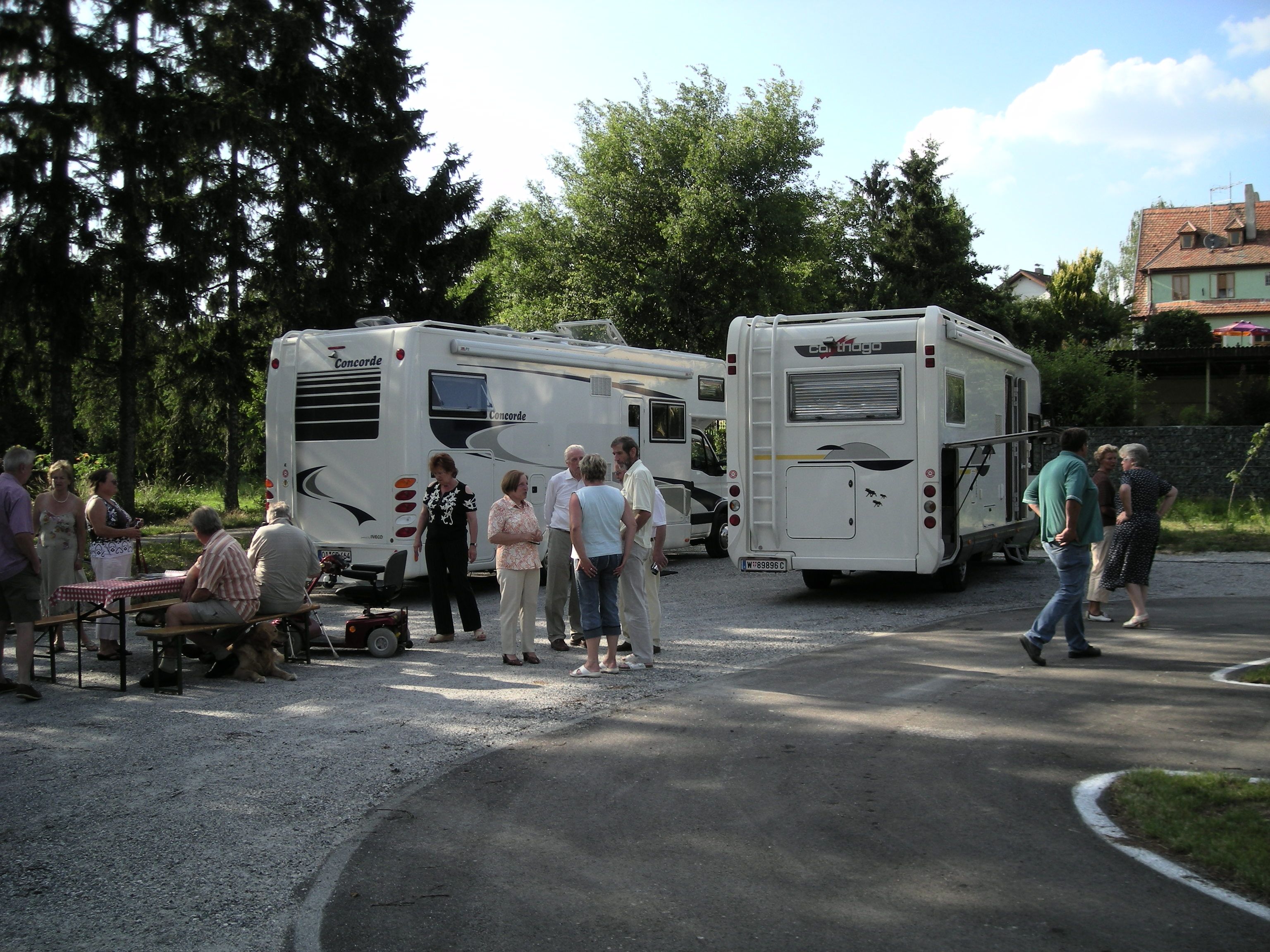 Lidé stojí a sedí vedle dvou mobilních domků na venkovním hřišti.