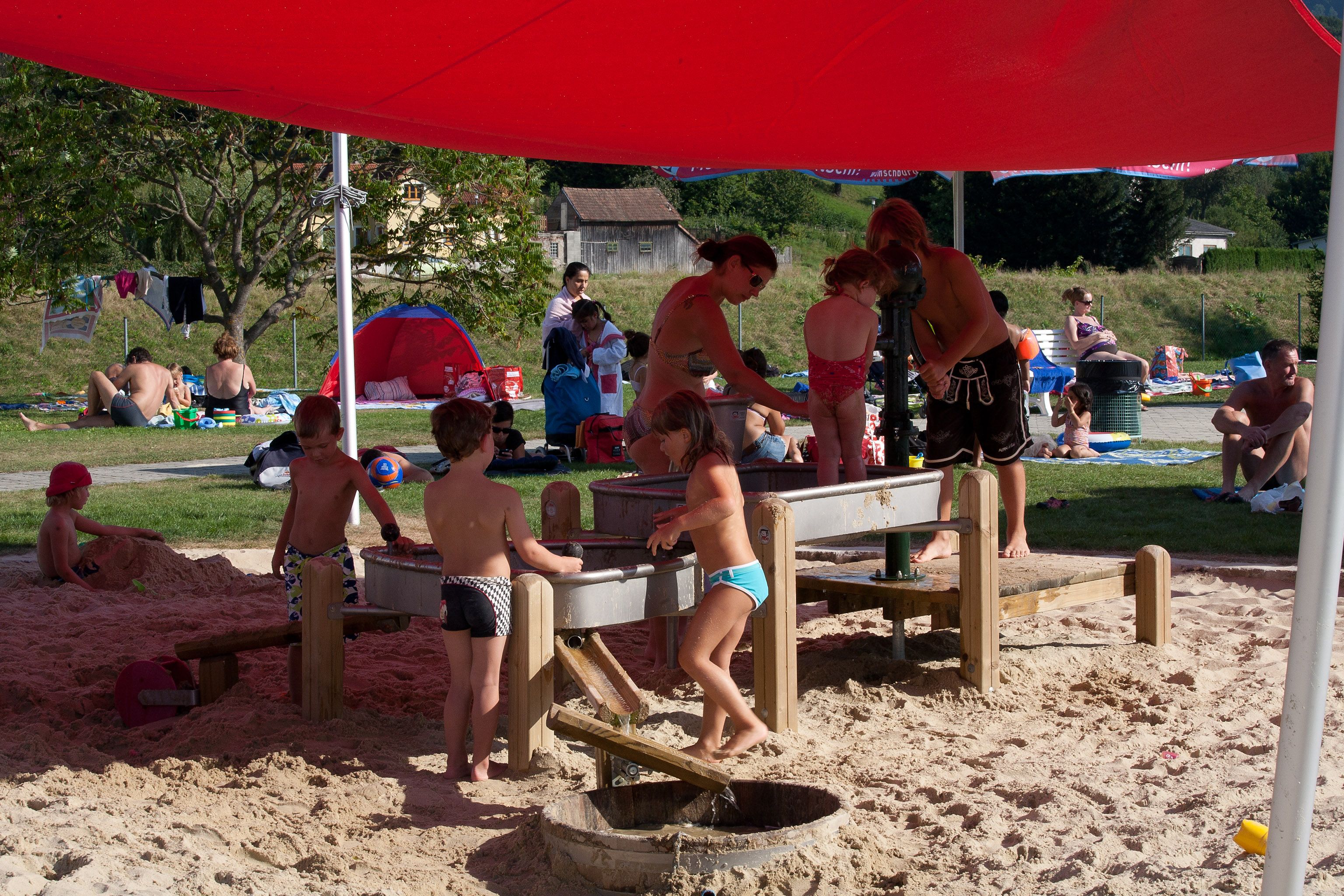 Děti si hrají v písku pod červenou markýzou ve Wilhelmsburg Parkbad.