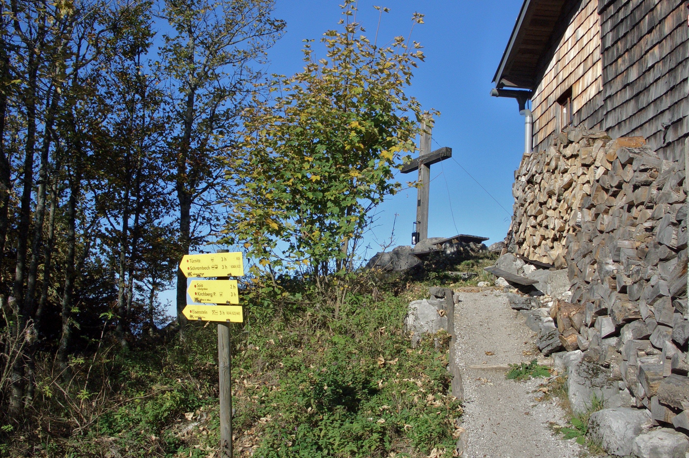 Rozcestník a dřevěný kříž vedle dřevěného domu v horské krajině.