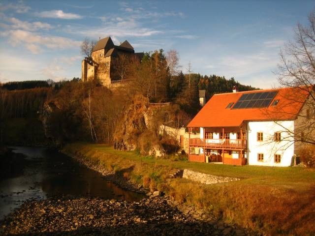 Hostinec s červenou střechou a solárními panely u řeky, s hradem na kopci v pozadí.