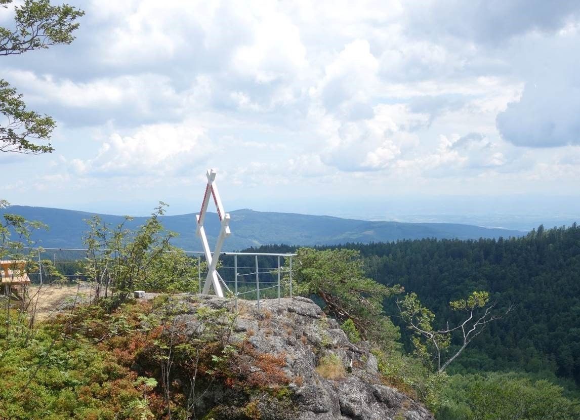 Vyhlídka na hoře s bílým rámem ve tvaru kříže a výhledem na zalesněné kopce pod zataženou oblohou.