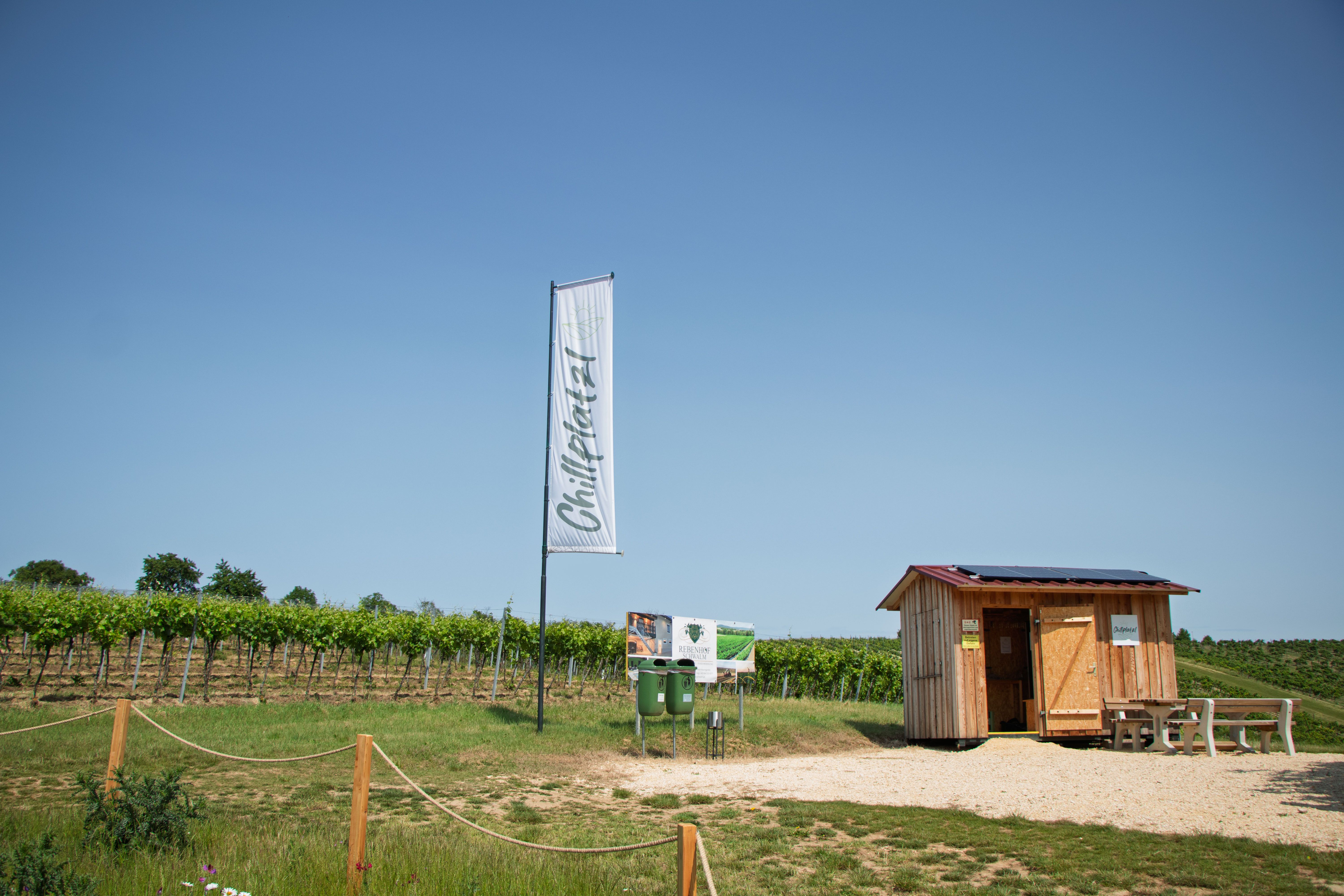 Malá dřevěná bouda vedle vinice s cedulí a vlajkou s nápisem „Chillplatz“.