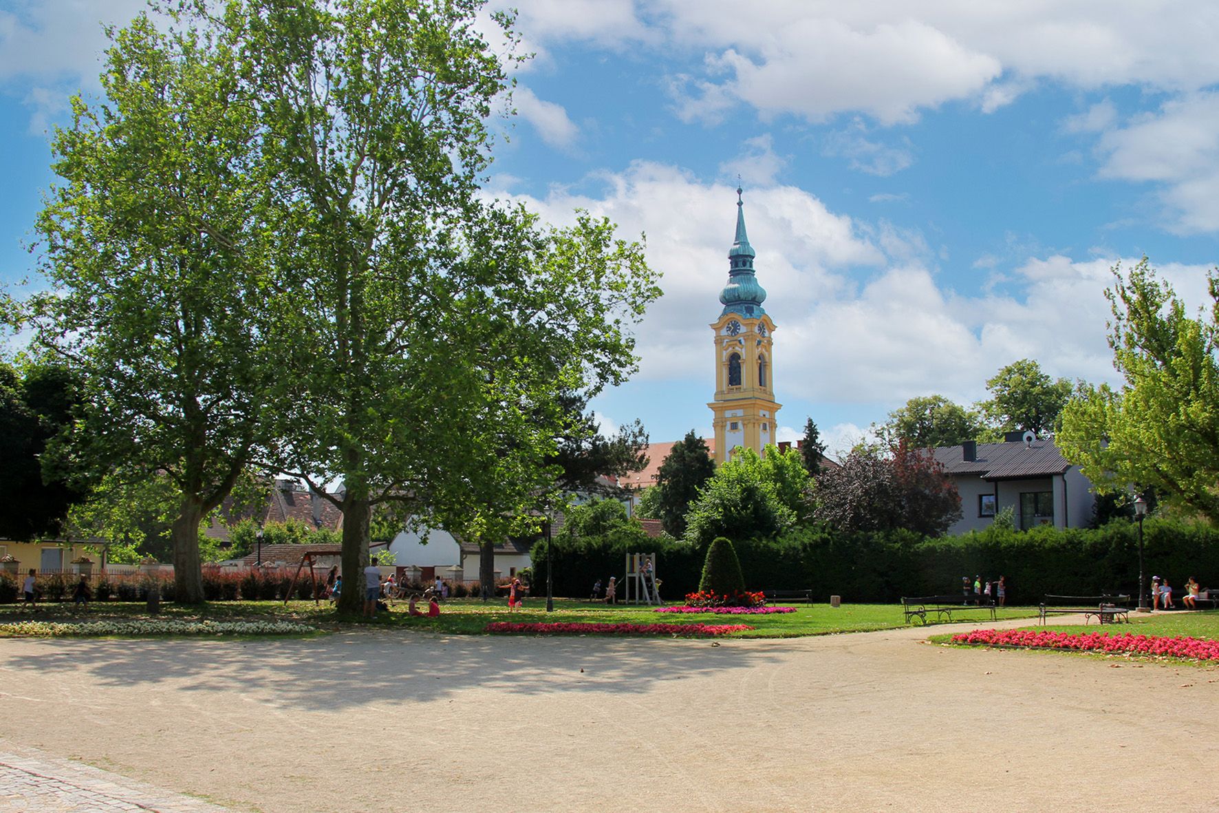 Park se stromy, květinovými záhony a kostelní věží v pozadí.