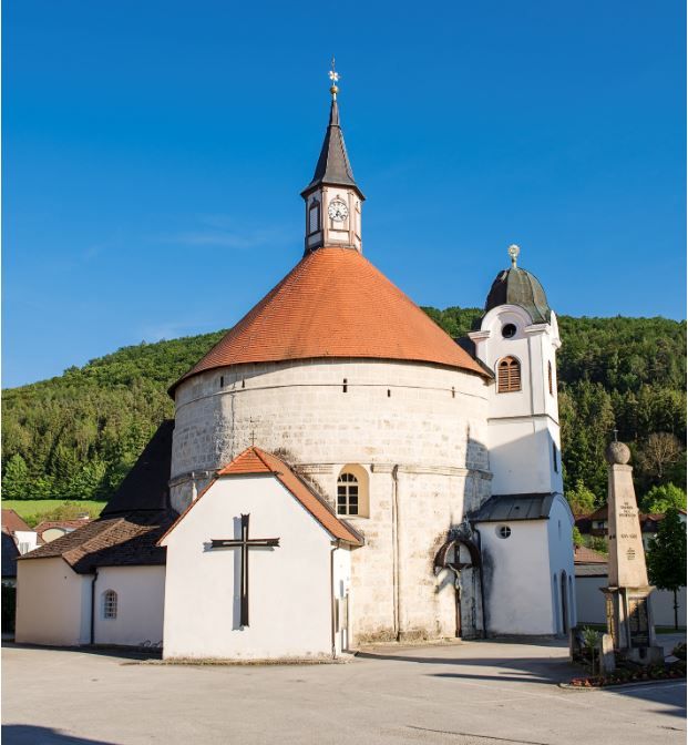 Farní kostel v Scheiblingkirchenu s kulatou věží a červenou střechou na pozadí modré oblohy.