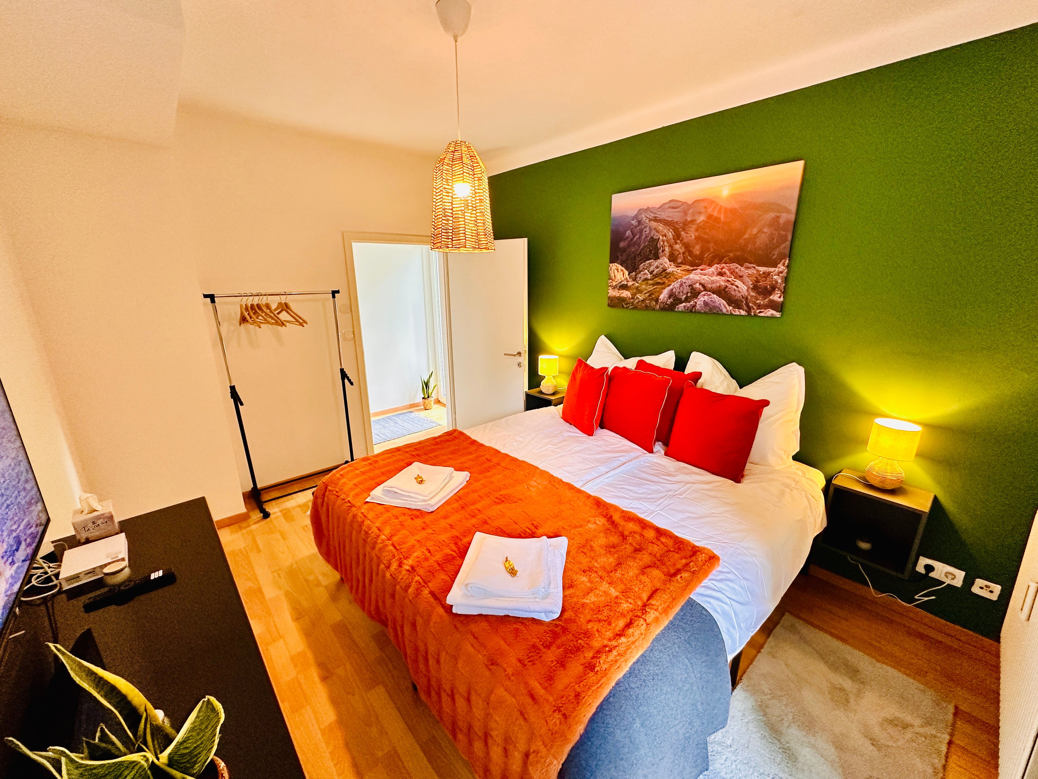 Útulná ložnice se zeleně natřenou stěnou, manželskou postelí s červenými polštáři, nočními stolky a lampou.